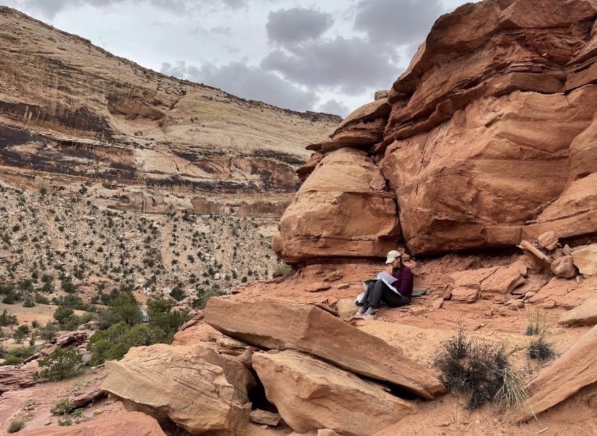 A person wearing a white hat, maroon shirt, and black pants sitting on red rock formations in a desert landscape with cliffs in the background.