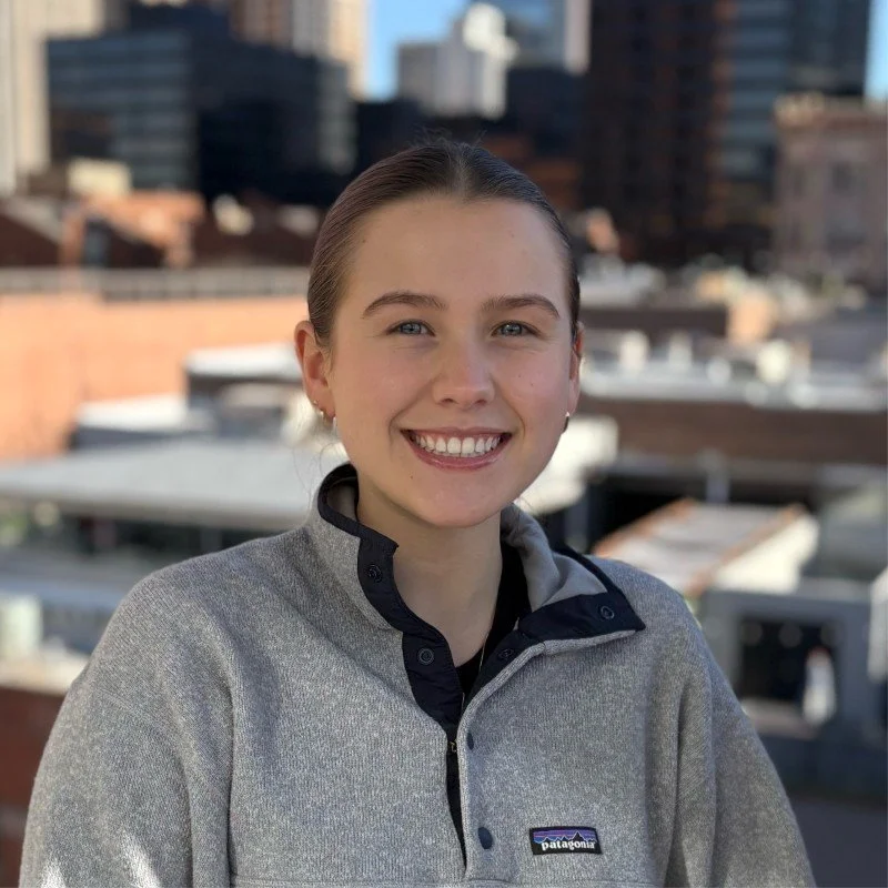 Young woman smiling outdoors in an urban setting, wearing a gray Patagonia jacket with city buildings in the background.
