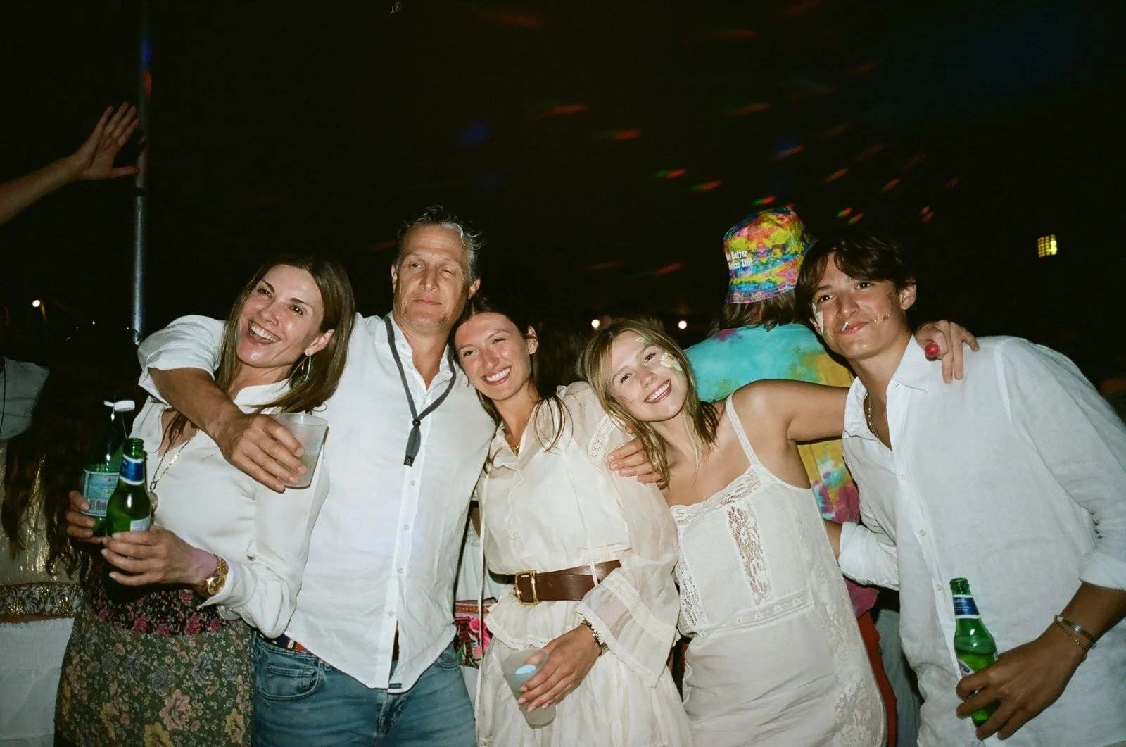 Group of six people smiling and enjoying a night event, some holding drinks, embracing each other, with dark background and colorful lights.