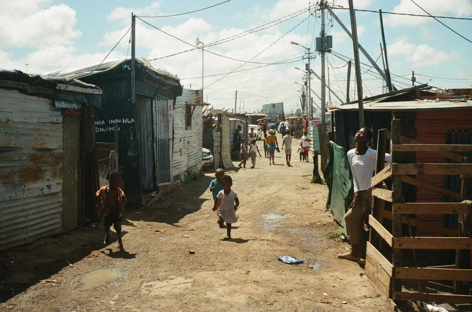 A dirt street in a neighborhood with makeshift houses made of metal sheets and wood, with children and adults walking and standing. There are overhead power lines and some small puddles on the ground.