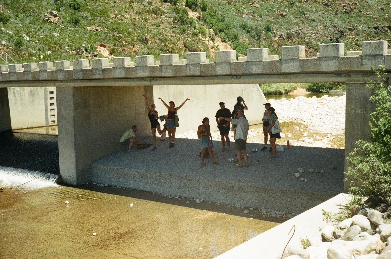 A group of people standing and sitting under a bridge over a river, with some rocks on the riverbank, near a hillside with green plants.