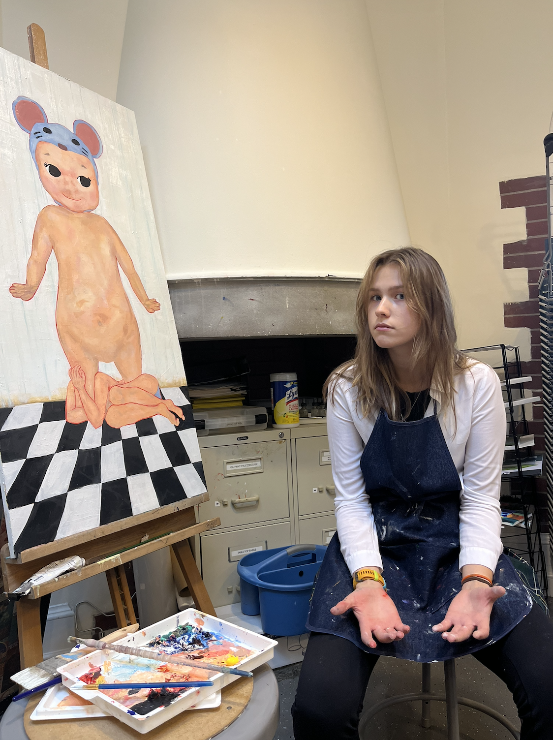 An artist sitting in her studio with a partially finished painting of a cartoonish nude female figure with a blue hat, kneeling on a checkered floor. Artist's supplies are visible around her.