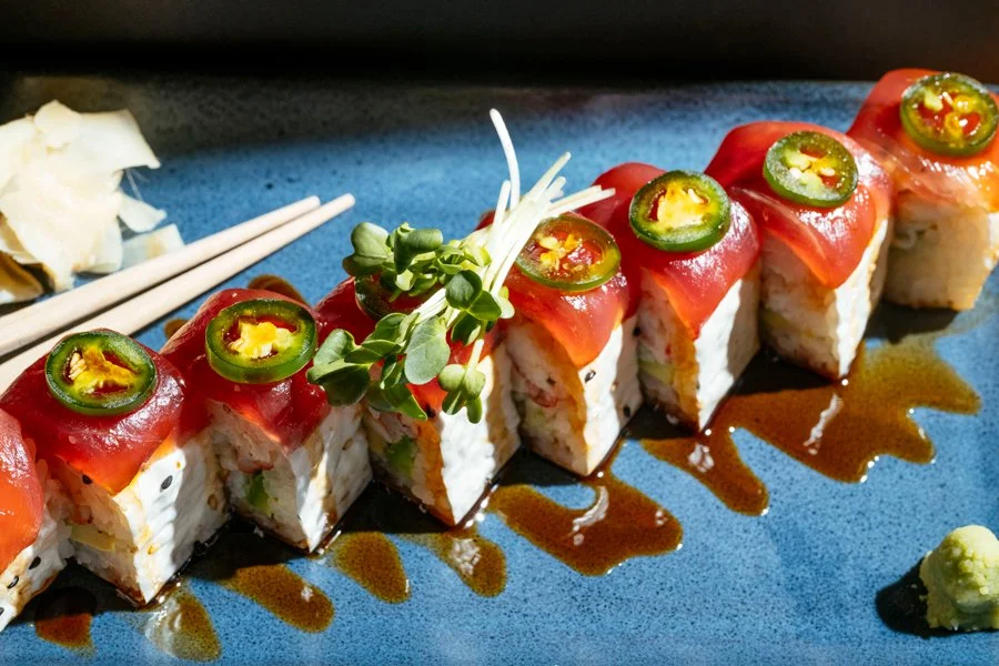 Sushi rolls topped with sliced jalapeños, salmon, and a drizzle of soy sauce, garnished with microgreens on a blue plate.