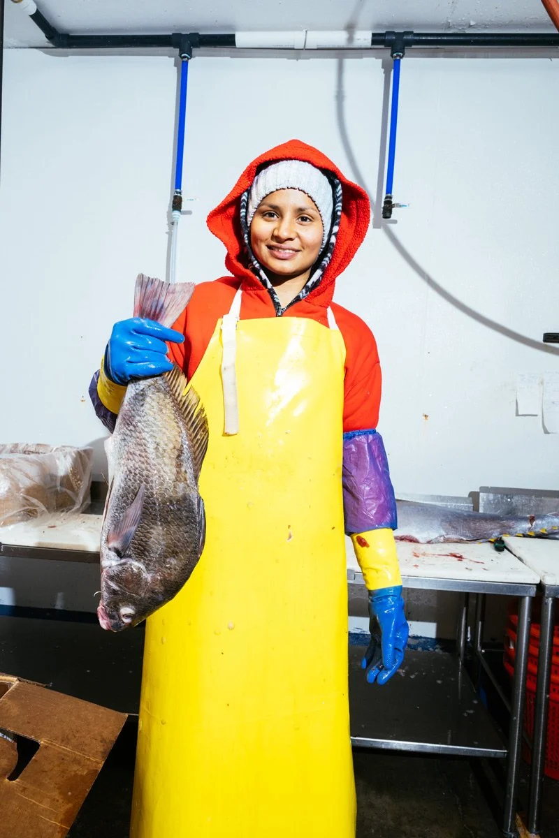 A woman in waterproof clothing and gloves holding a large fish inside a fish market or processing facility.