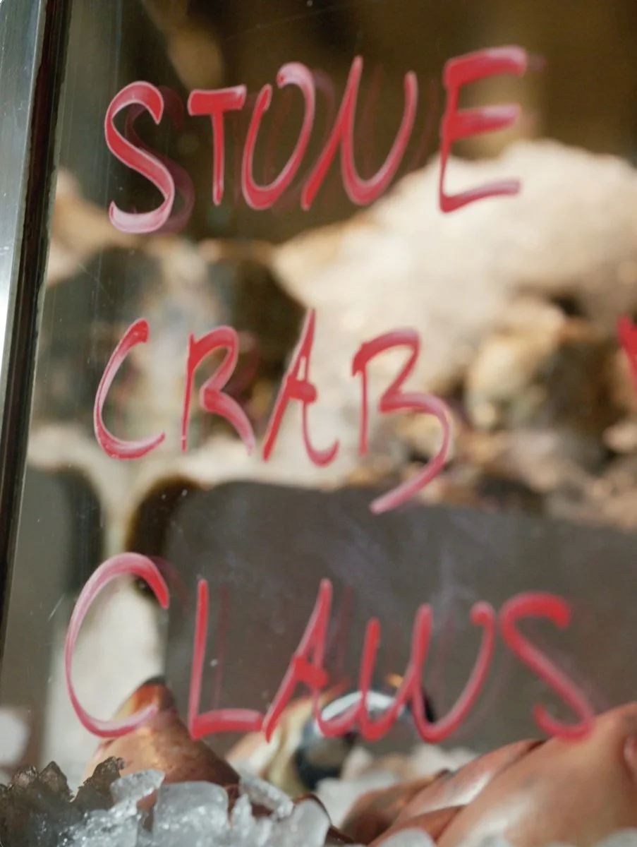 A glass display case with a handwritten sign in red and pink marker that reads 'STONE CRAB CLAWS' in front of some stone crab claws and ice.