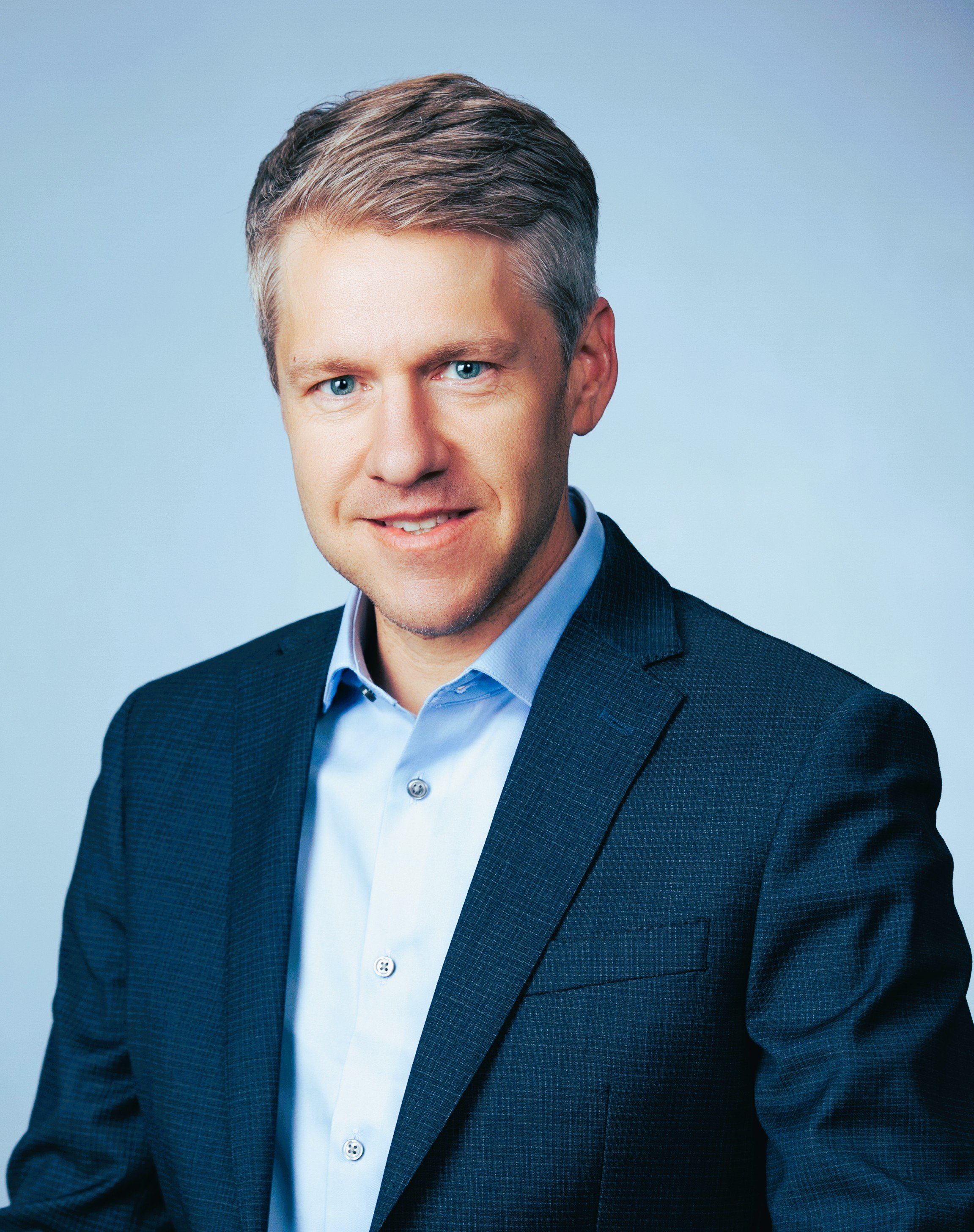 Professional headshot of a smiling man in a dark blue suit and light blue shirt against a pale blue background