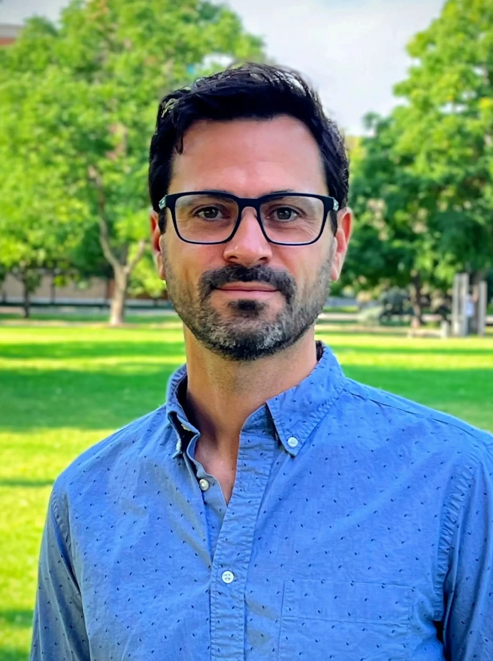 Portrait of a man wearing glasses and a blue button-down shirt outdoors in a sunny park