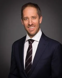 Professional headshot of a smiling man in a dark suit, white shirt, and striped tie against a gray background.