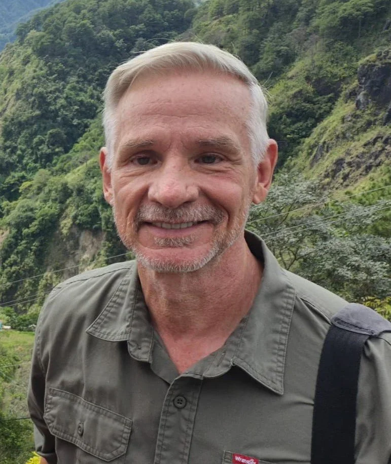 Smiling gray-haired man wearing an olive Wrangler shirt and backpack in front of a lush mountain landscape