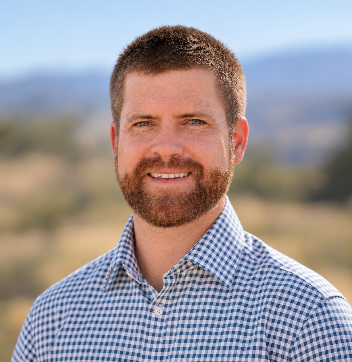 Smiling bearded man in a blue checkered shirt outdoors with a blurred landscape background
