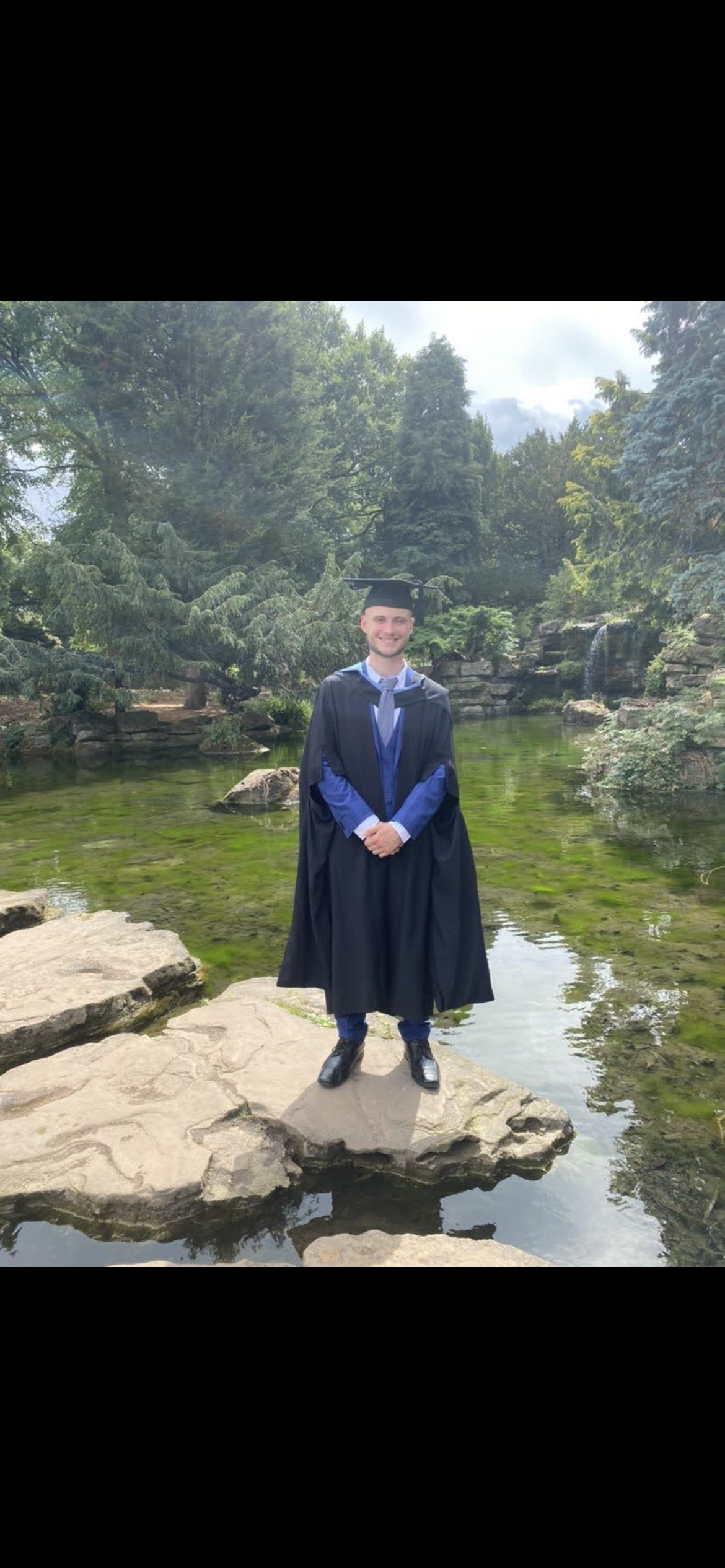 Graduate in cap and gown standing on rocks beside a landscaped pond and waterfall