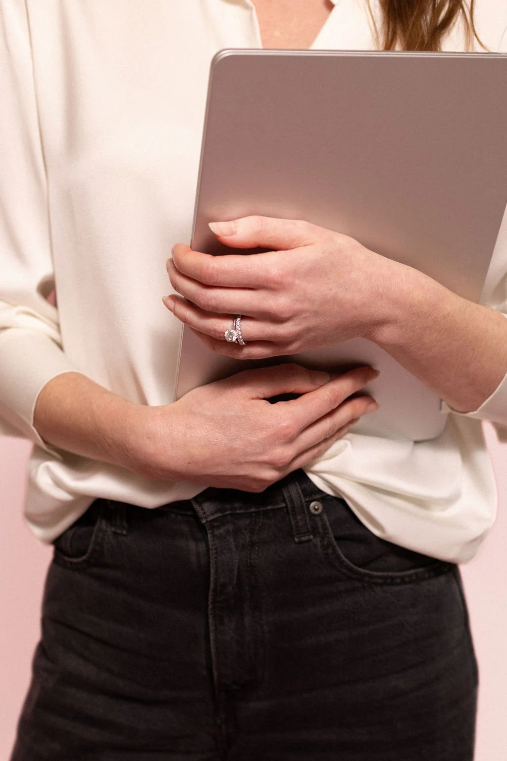 Close-up of a woman holding a laptop with both hands, wearing a white blouse and black jeans, with a diamond ring on her finger.