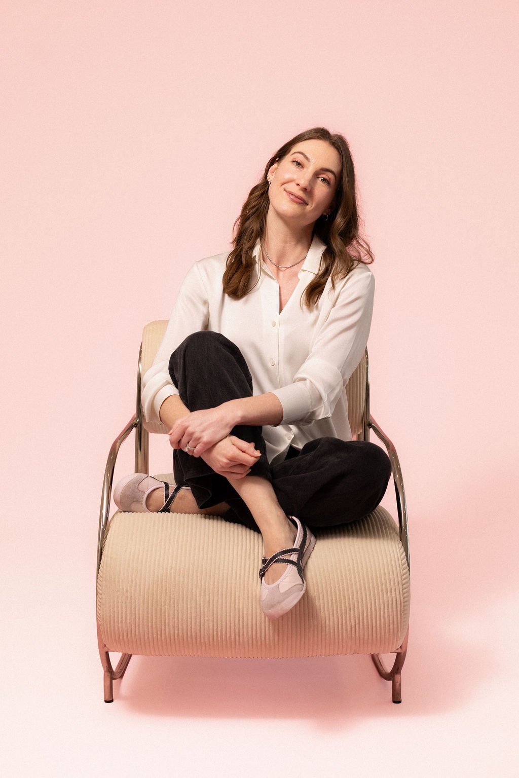 A woman with long brown hair, sitting on a beige chair with a metal frame against a light pink background, smiling gently with her head tilted.