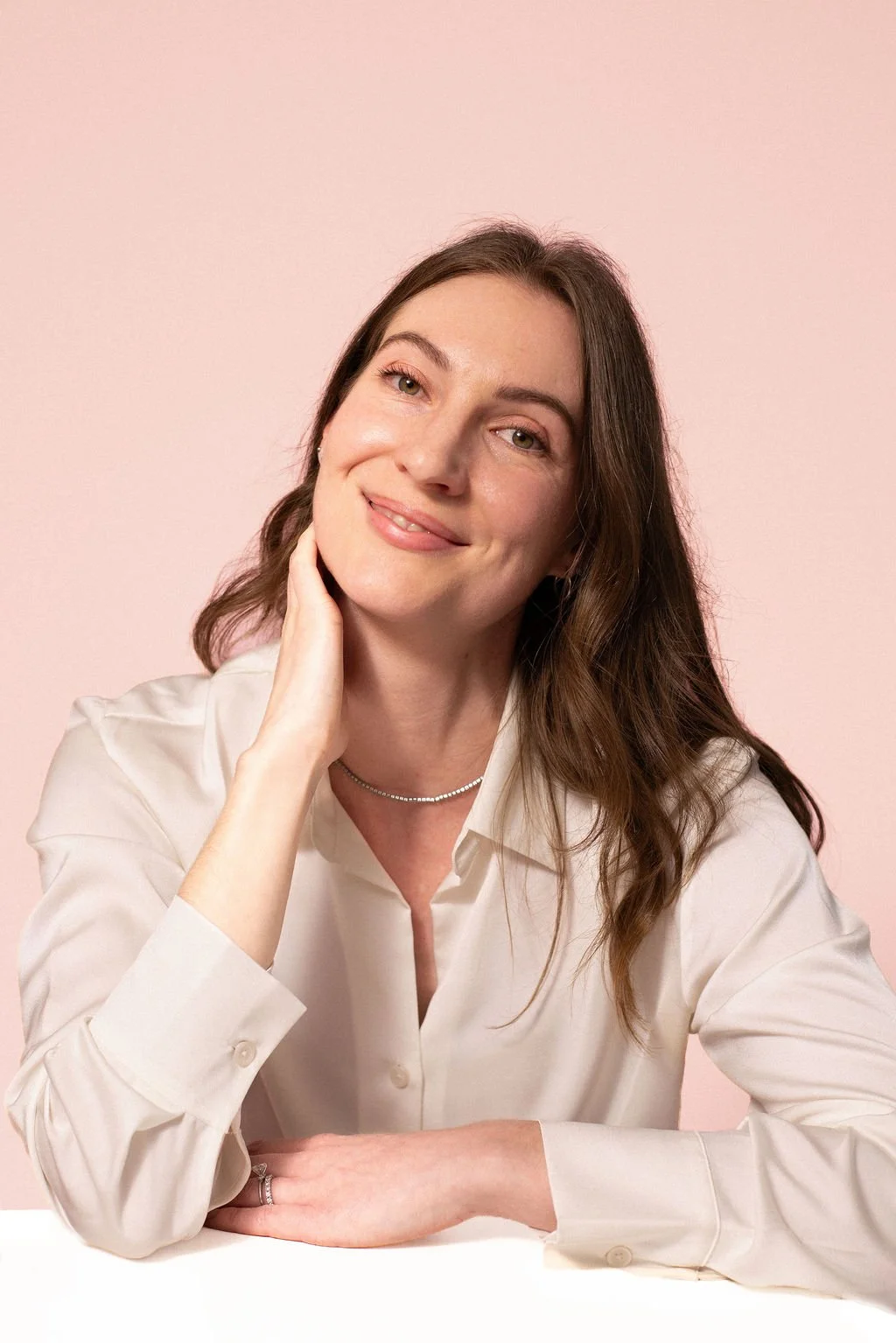 A young woman with long brown hair, smiling, wearing a cream-coloured shirt and tennis necklace, posed against a pink background.