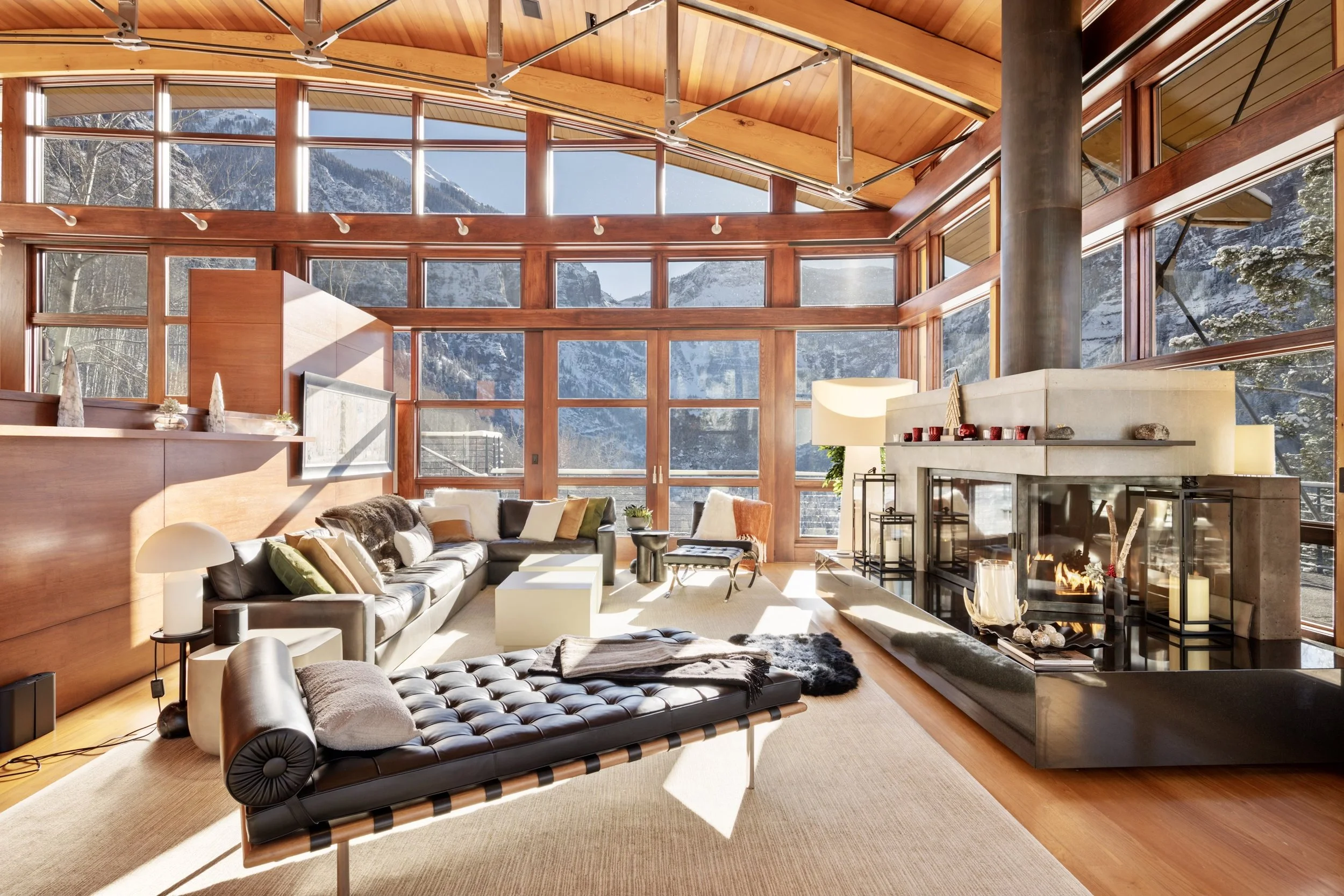 Living room with large windows showing snowy mountain landscape, wooden ceiling and walls, modern fireplace, beige carpet, black leather lounge chair, white side table, colorful pillows on a sofa, and various decorative items.