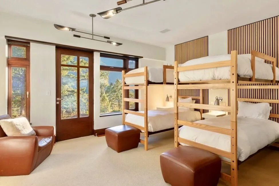Room with four bunk beds, two leather armchairs, two leather ottomans, large windows, and a door leading outside, decorated in a modern style with wooden accents and natural light.