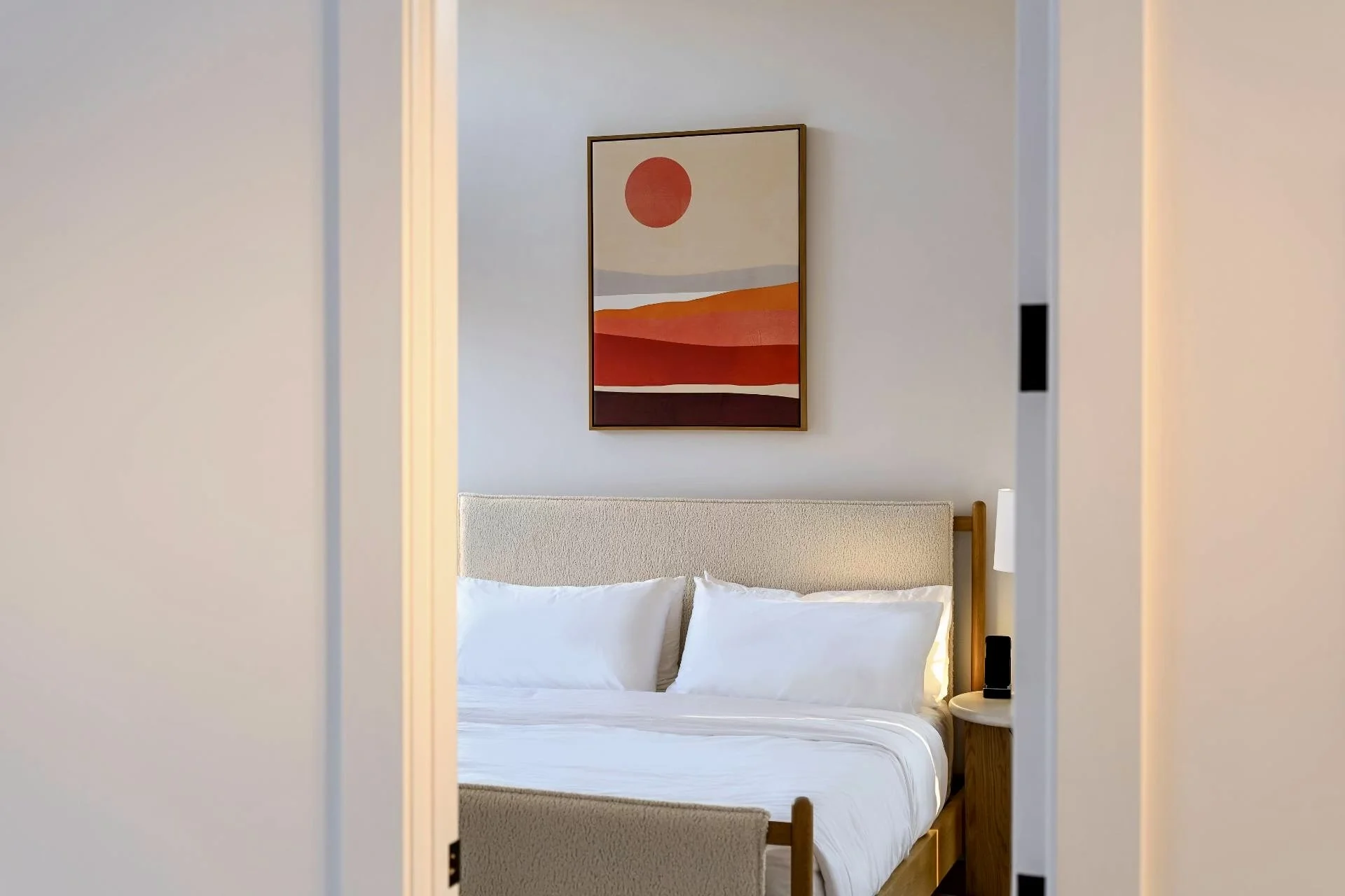 A bedroom viewed through a doorway shows a bed with white bedding, a beige headboard, a nightstand with a lamp, and a framed abstract landscape painting with shades of red, orange, gray, and white on the wall.