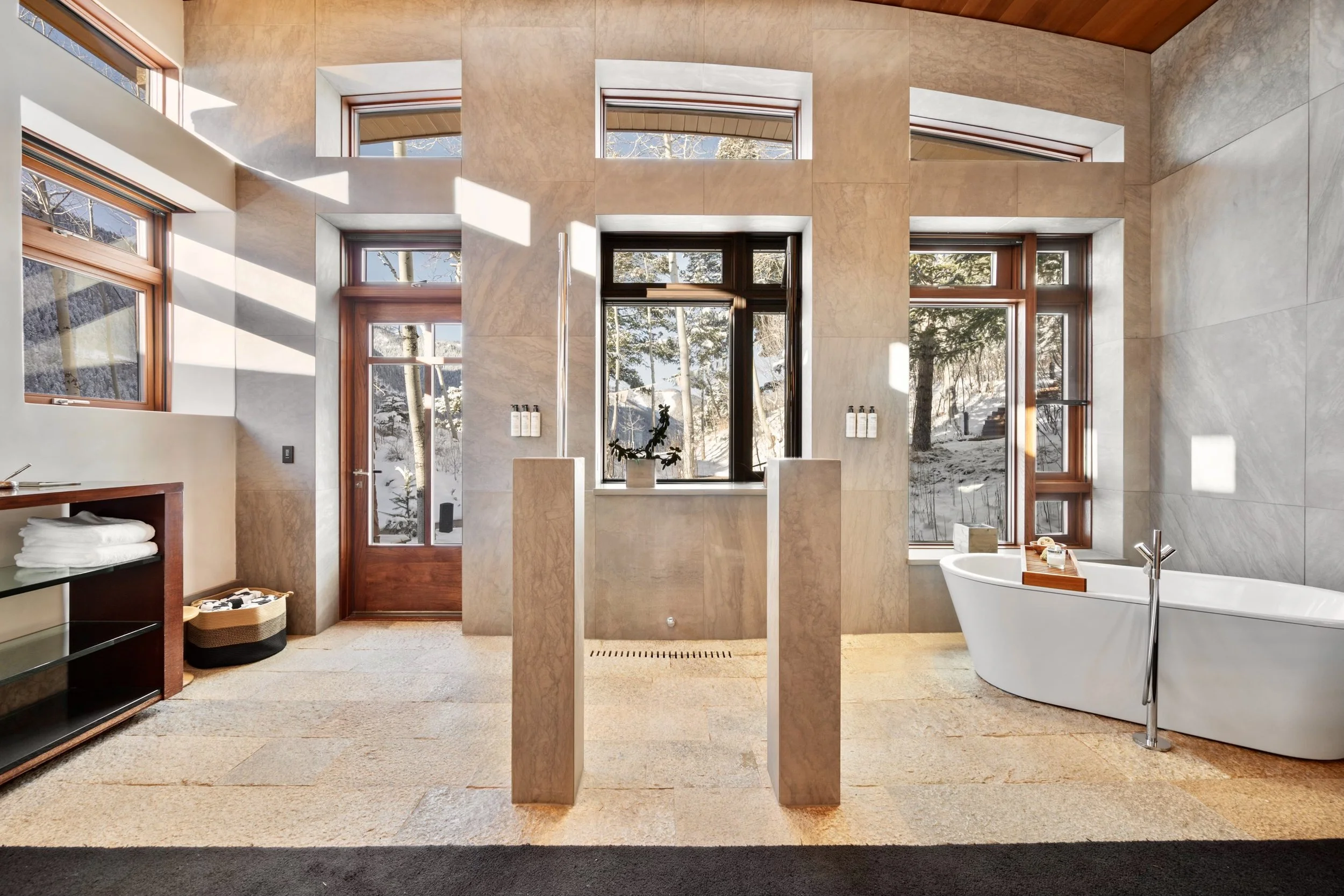 A modern bathroom with large windows showing a snowy outdoor landscape, a freestanding bathtub, a potted plant on the windowsill, and beige tiled walls.
