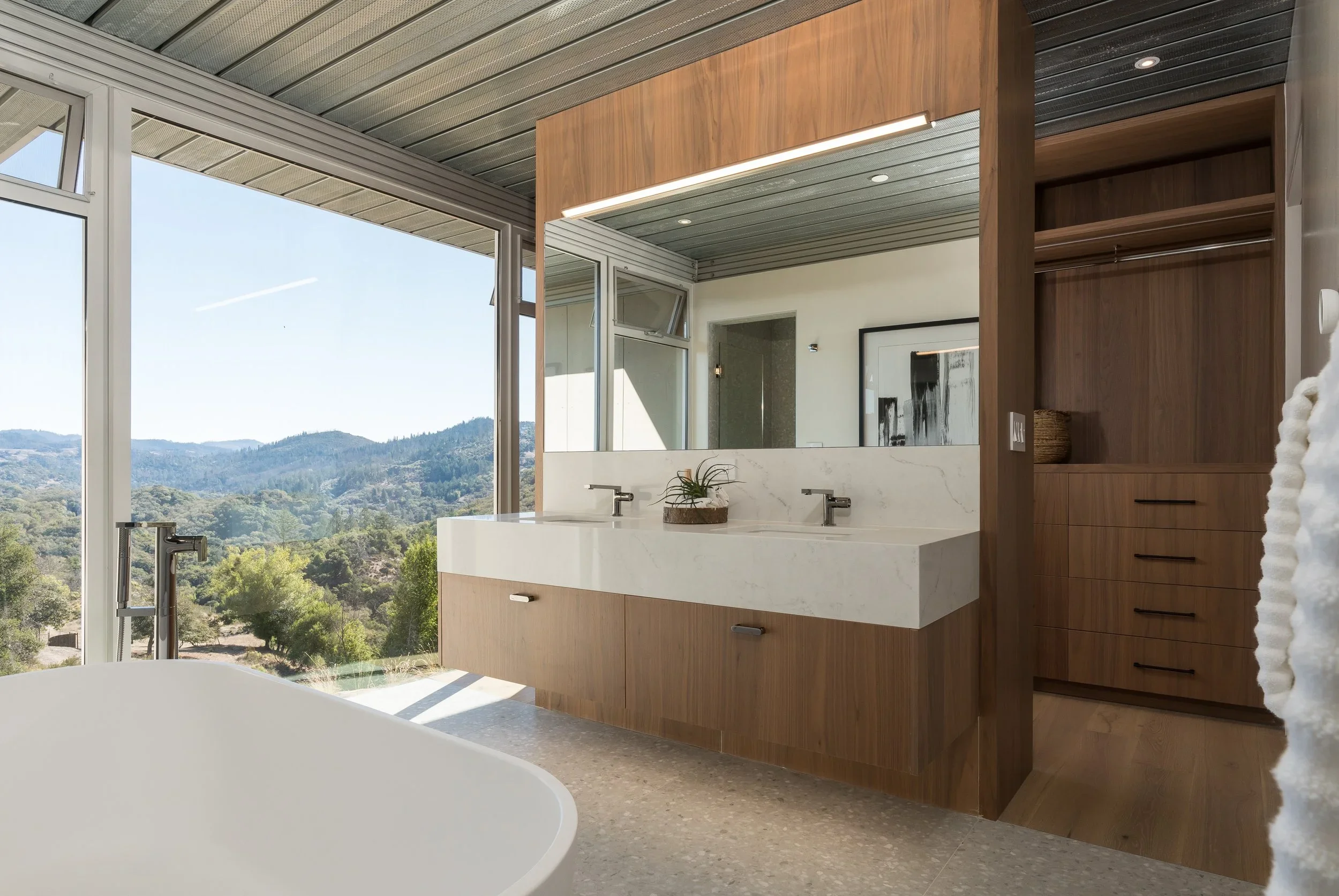 Modern bathroom with large windows showing a scenic mountain view, featuring a white bathtub, wooden vanity with a marble countertop, dual sinks, and a wooden cabinet, with a textured ceiling and a potted plant on the countertop.