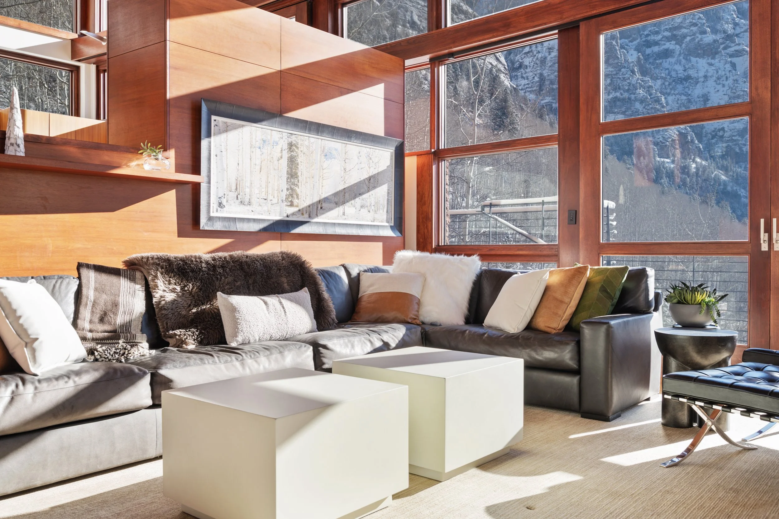 Living room with large floor-to-ceiling windows showing snowy mountain landscape, a large gray sectional sofa with multiple pillows, white cube-shaped coffee tables, and a black ottoman.