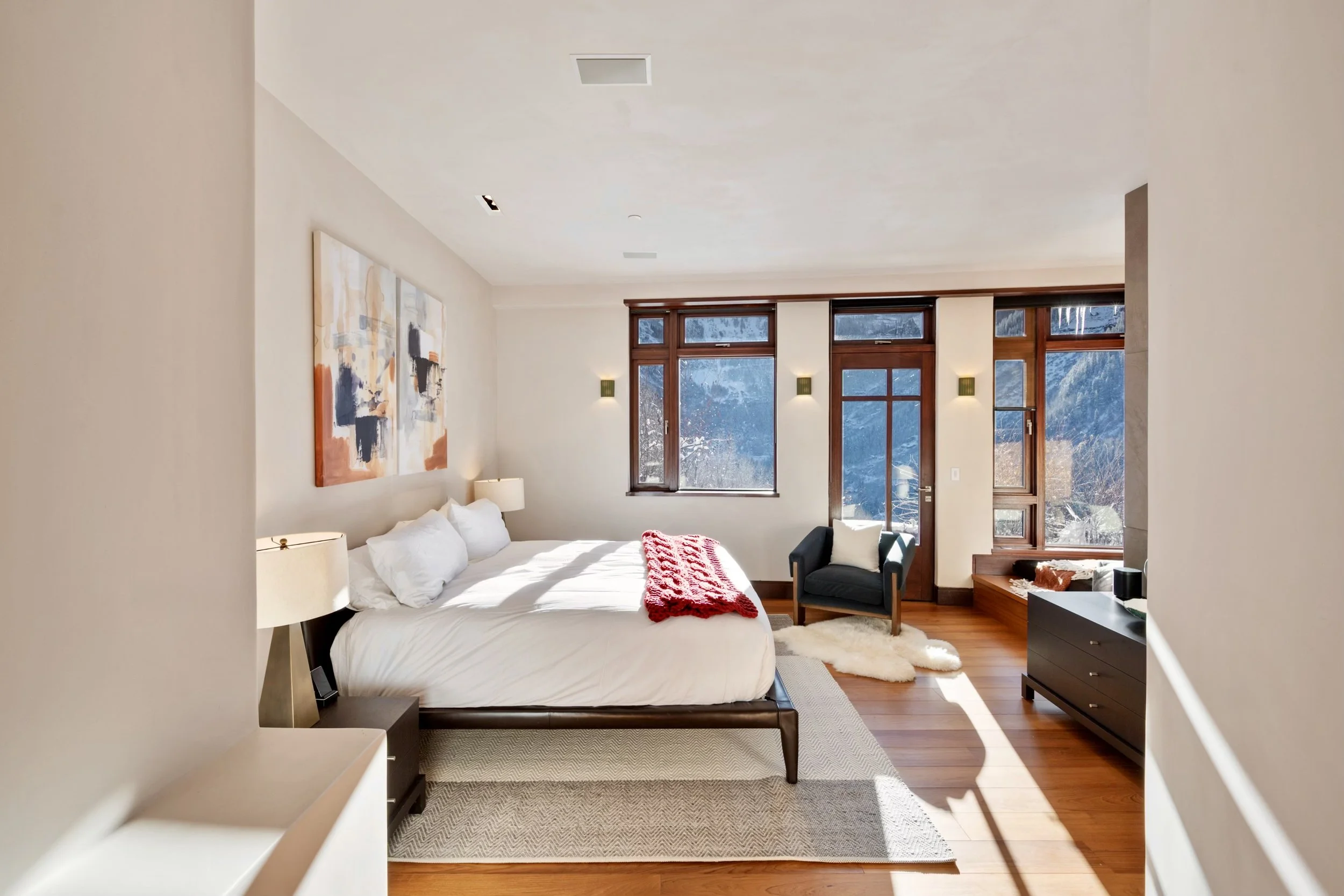 A bedroom with a bed, artwork on the wall, a nightstand with a lamp, large windows showing snowy mountains outside, and hardwood floors.