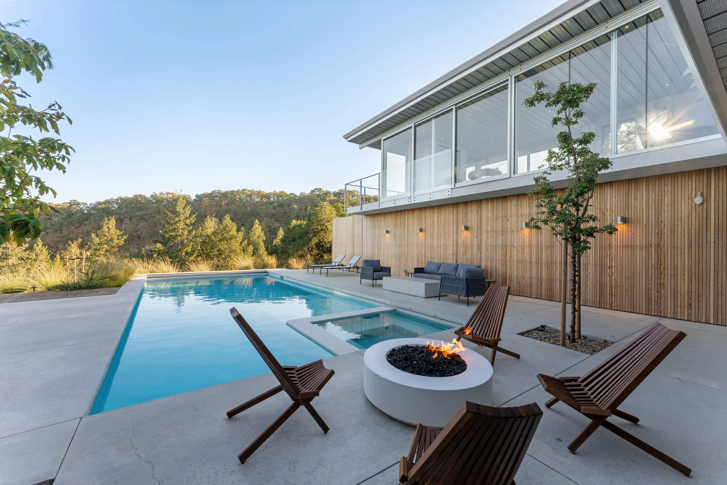 Modern house with large glass windows, outdoor pool, patio furniture, and a fire pit, surrounded by trees and hills under a clear blue sky.