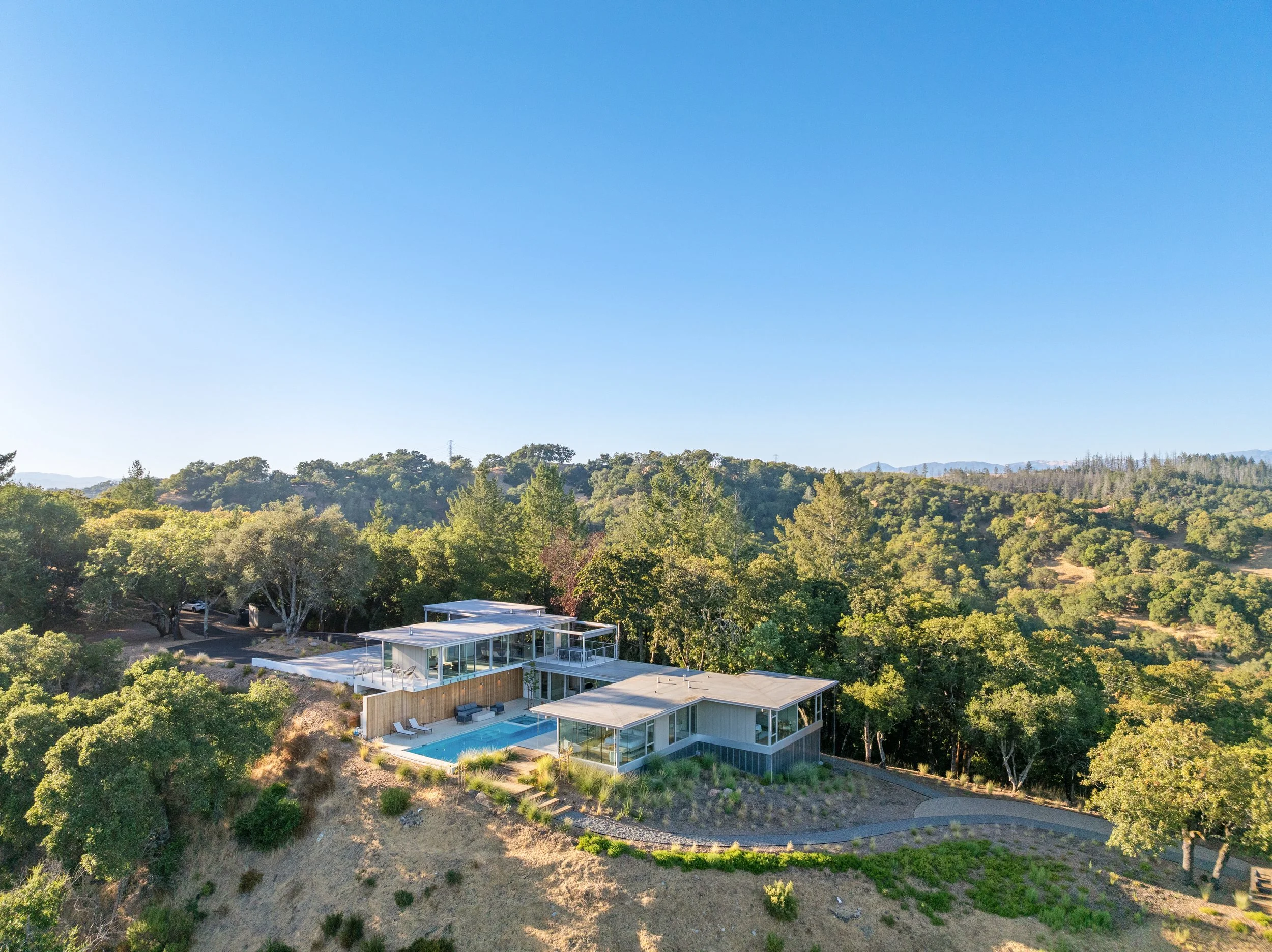 A modern house with large windows and a swimming pool, situated on a hillside surrounded by trees with dense forest and a clear blue sky in the background.