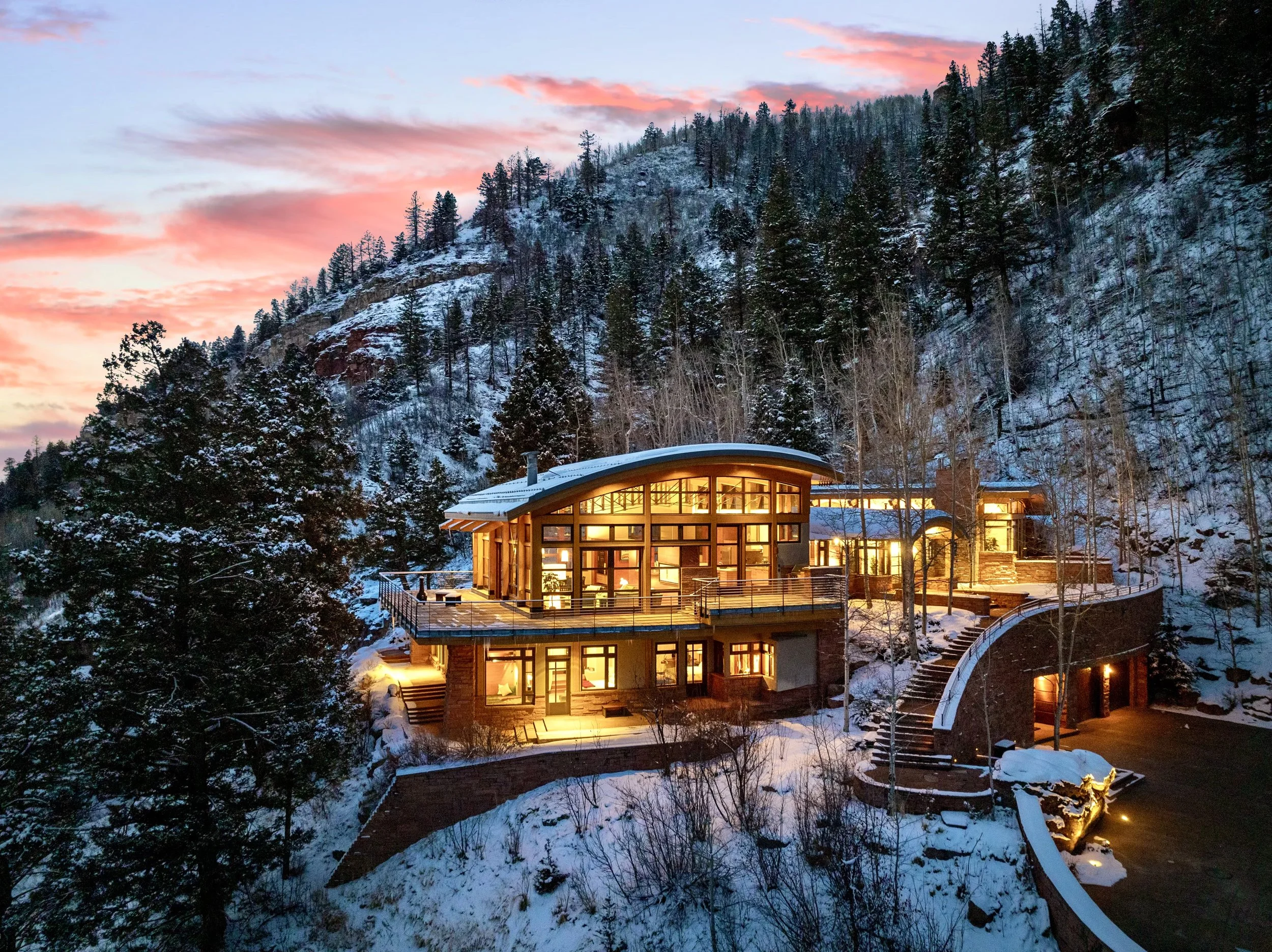 A house built into a snowy hillside with large windows illuminated from inside, surrounded by trees and a winding staircase at sunset.