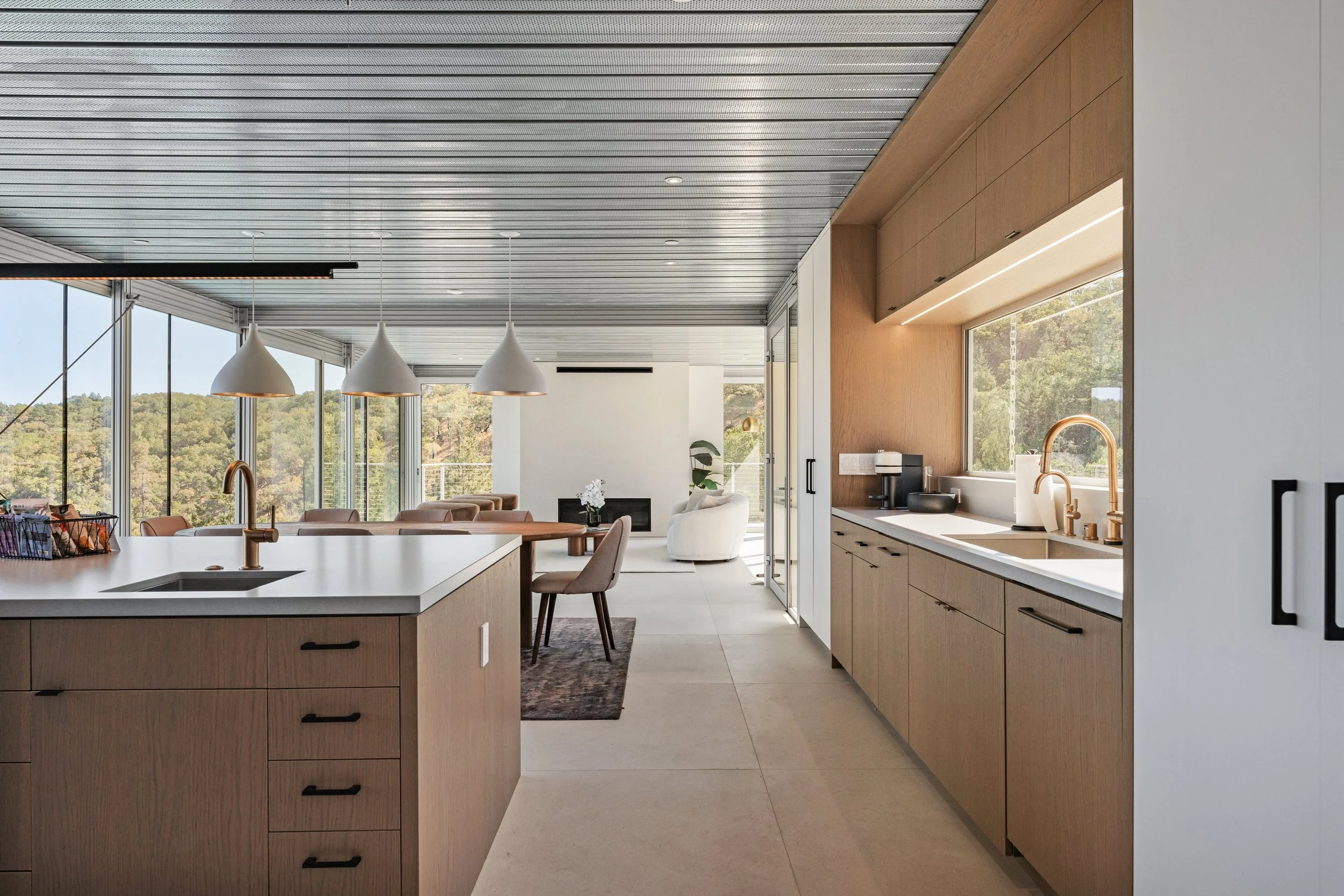 Modern kitchen and living room with large windows, wooden cabinets, white countertops, pendant lighting, and outdoor scenery.