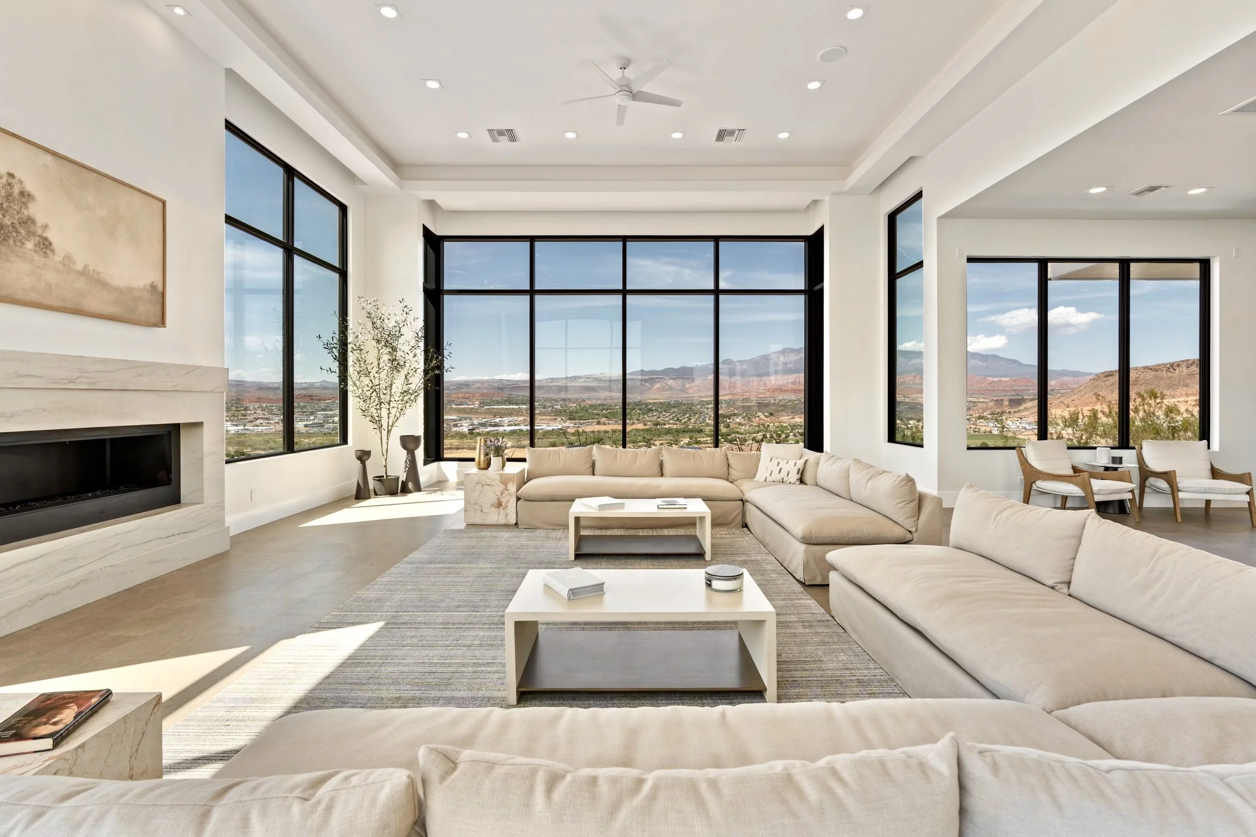 Modern living room with large windows showcasing a desert landscape and mountains in the background, white walls, beige sectional sofa, white coffee tables, and minimal decor.