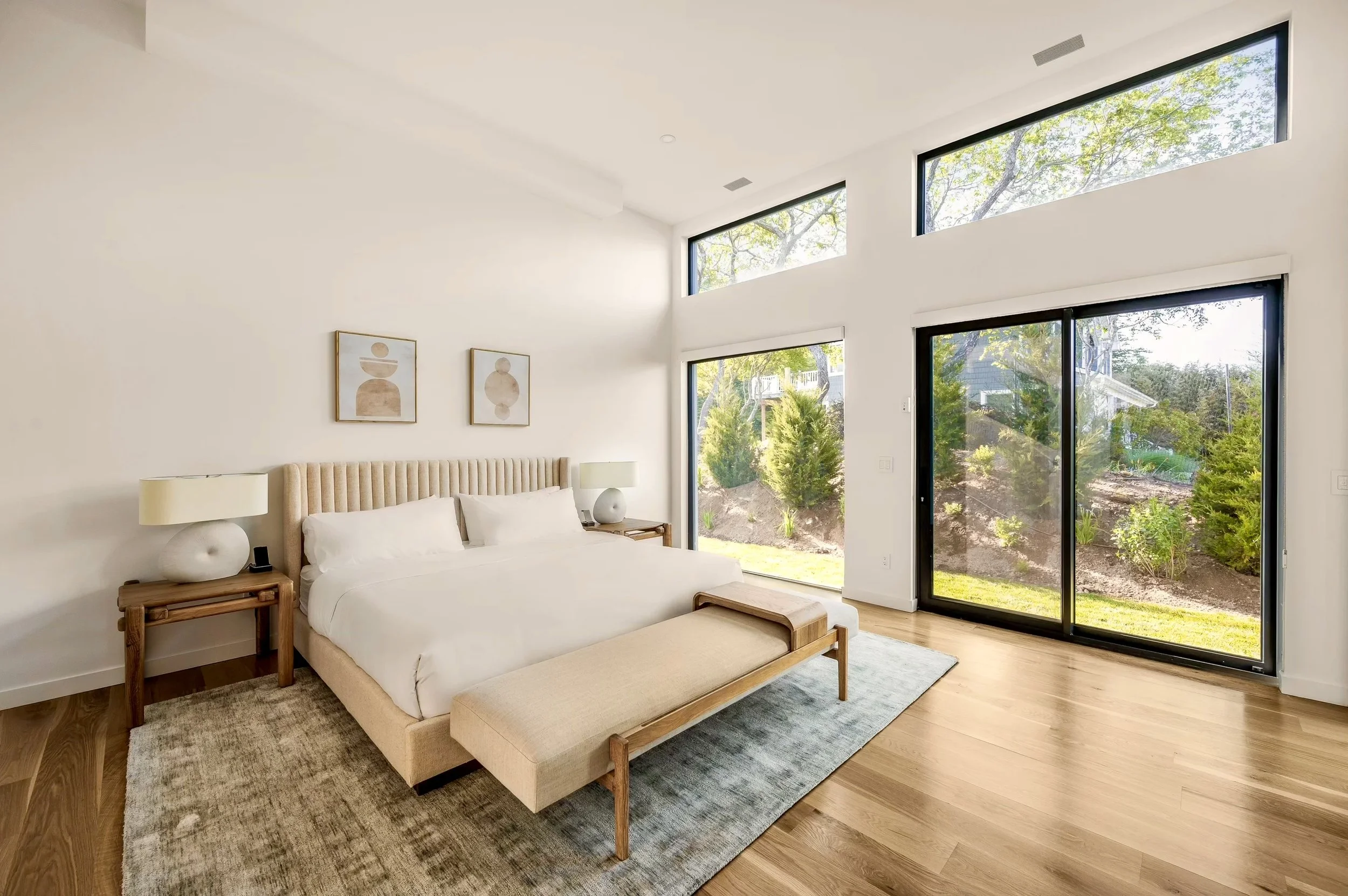 Modern bedroom with a beige upholstered bed, two white bedside lamps, large windows showing trees and greenery outside, minimalist artwork above the bed, wooden furniture, and light-colored hardwood floors.