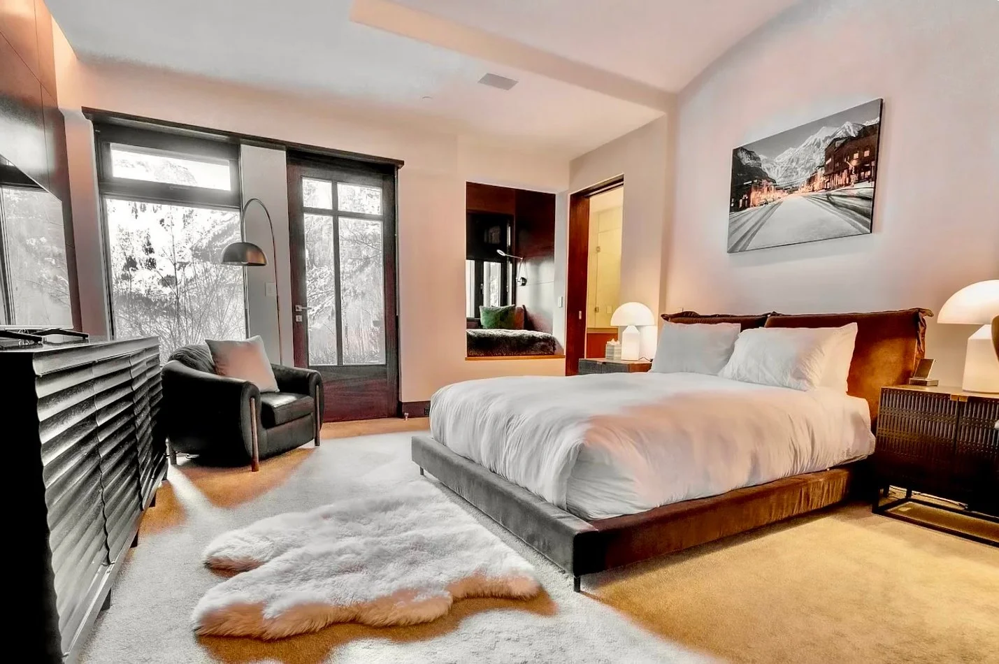 A modern bedroom featuring a large bed with white linens, wooden headboard, nightstands with lamps, a black armchair with a pillow near a glass door, and a fur rug on carpeted floor, with a window seat and framed mountain scenery photo on wall.
