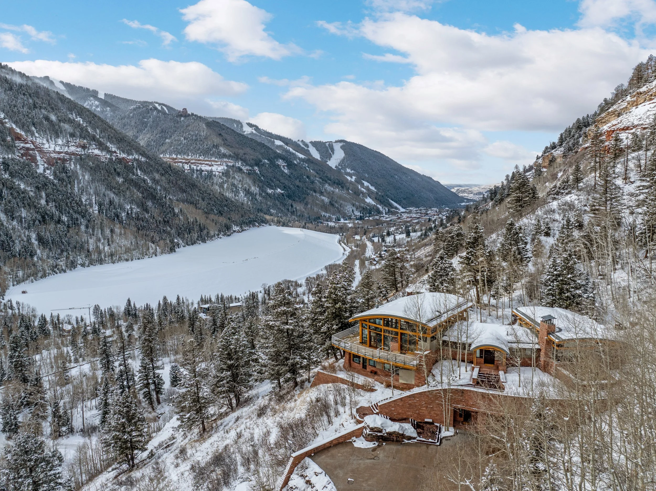 A snowy mountain landscape with dense forests, a frozen river or lake, and a modern building with large windows and a terrace in the foreground.