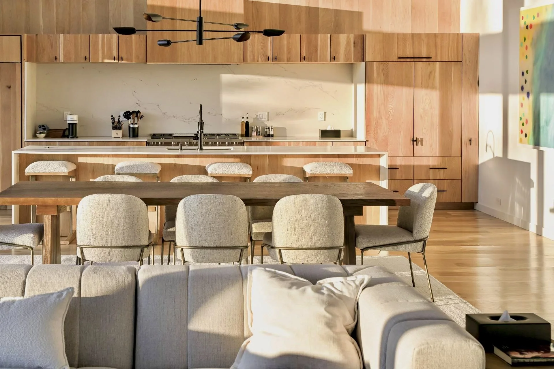 Open-concept kitchen and dining area with light wood cabinetry, white marble backsplash, and beige upholstered chairs around a wooden dining table. Sunlight filters into the space.