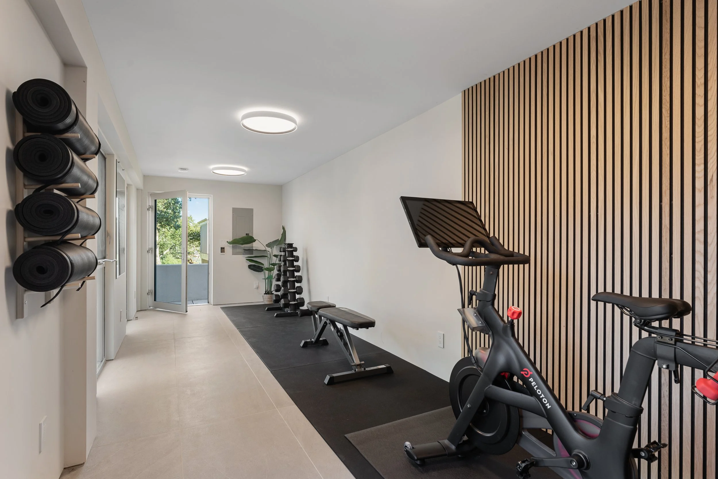 A modern home gym with exercise bikes, yoga mats, and a wall with wooden slats, near a door leading outside, natural light coming in.