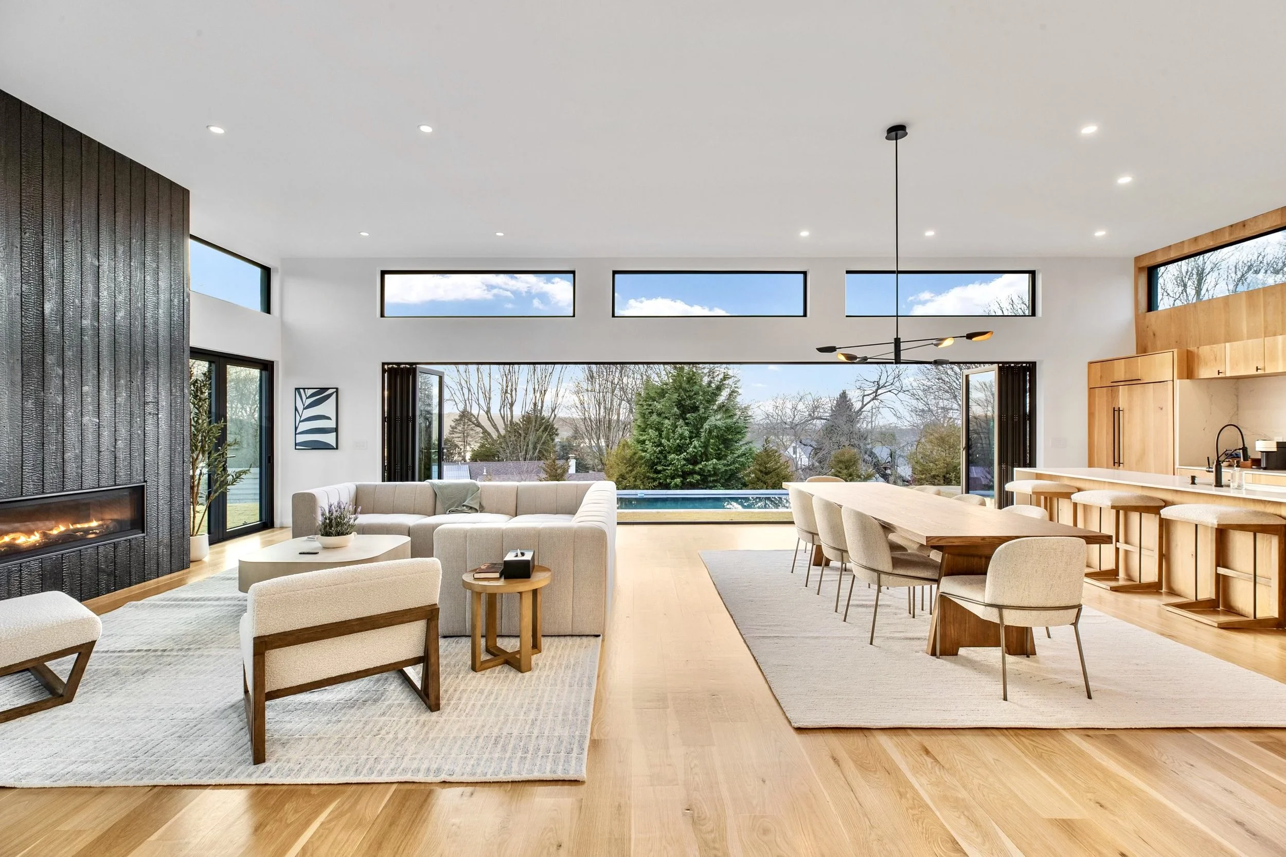 Modern open-concept living room with large windows showing trees and a sky, light wood flooring, white walls, a fireplace, beige sofa, wooden chairs, and a kitchen area with bar stools.