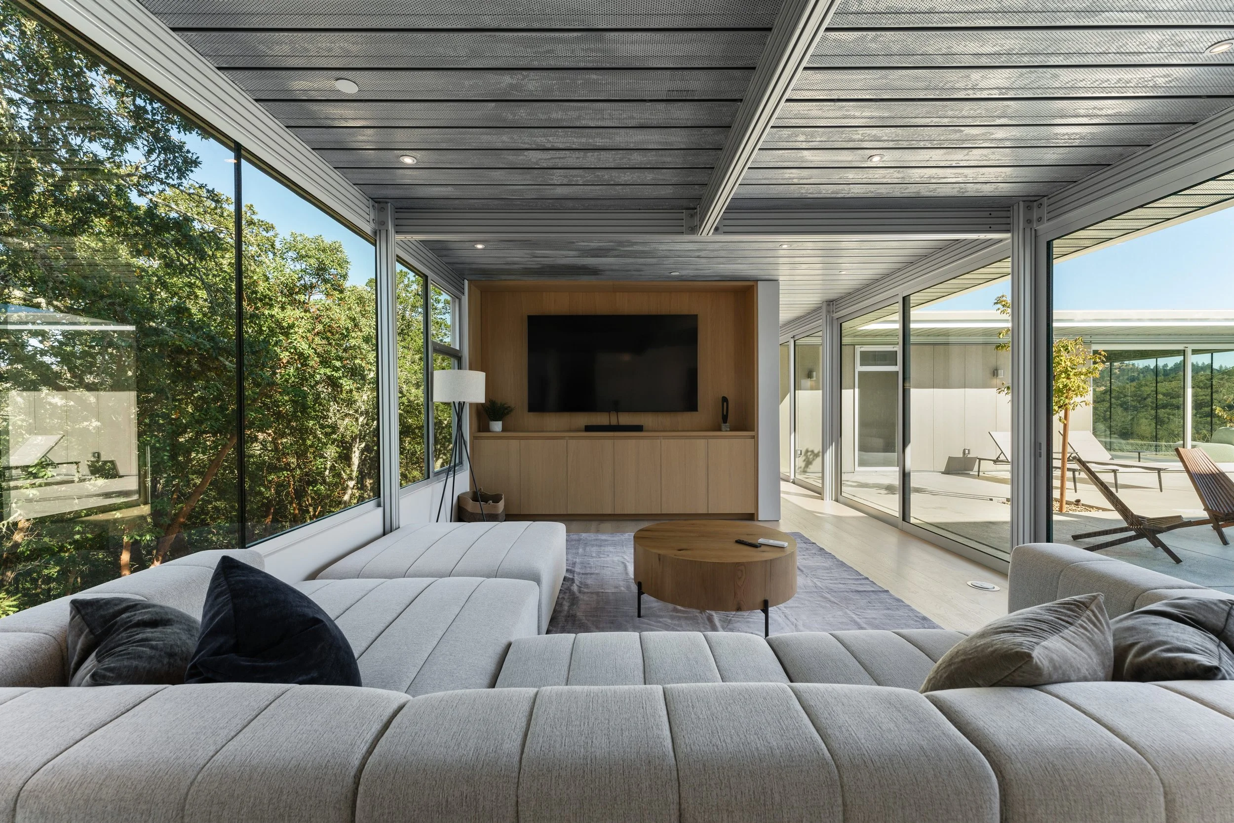 Modern living room with large glass windows and sliding doors, tan couch with black and gray pillows, wooden coffee table, TV mounted on a wooden accent wall, and outdoor lounge chairs visible through the windows.