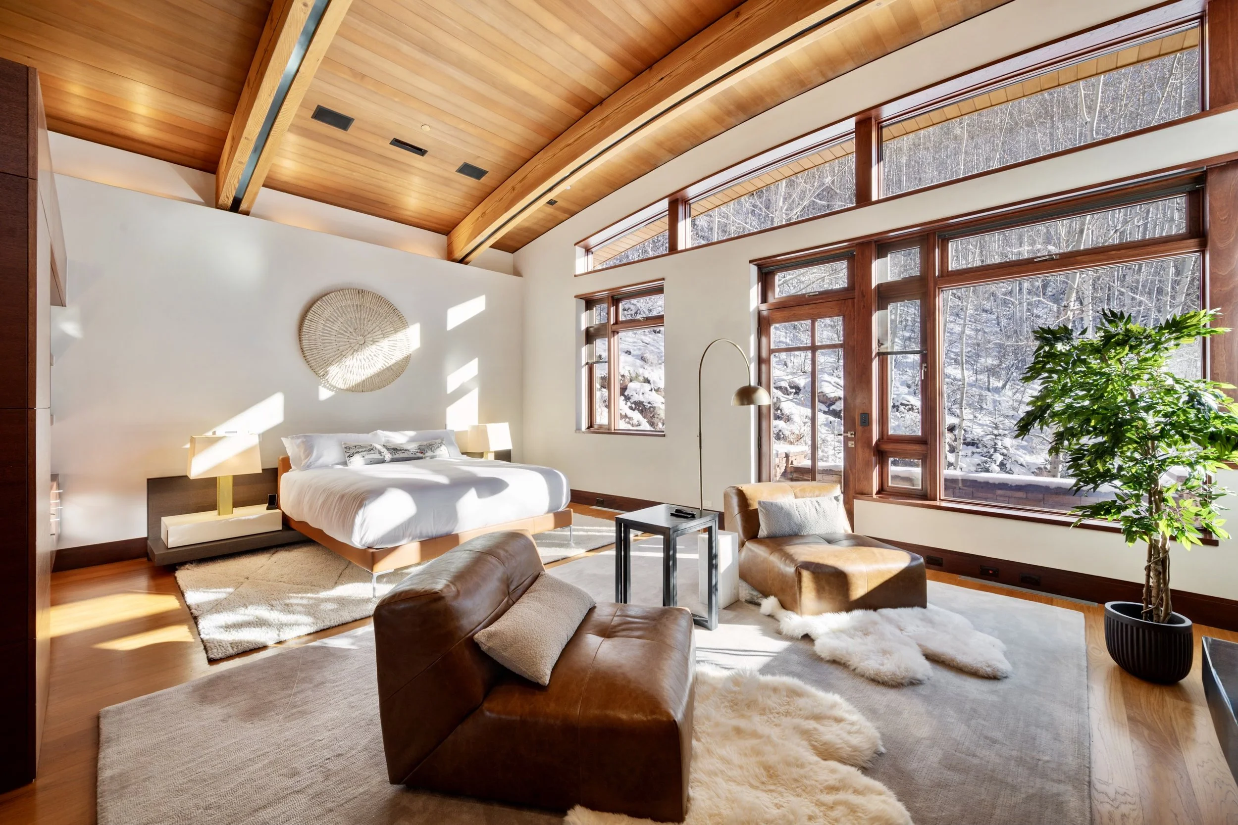 A spacious bedroom with large windows revealing snowy outdoor scenery. The room features a wood-paneled vaulted ceiling, a large bed with white bedding, a beige area rug, leather chairs, a modern floor lamp, and a potted plant.