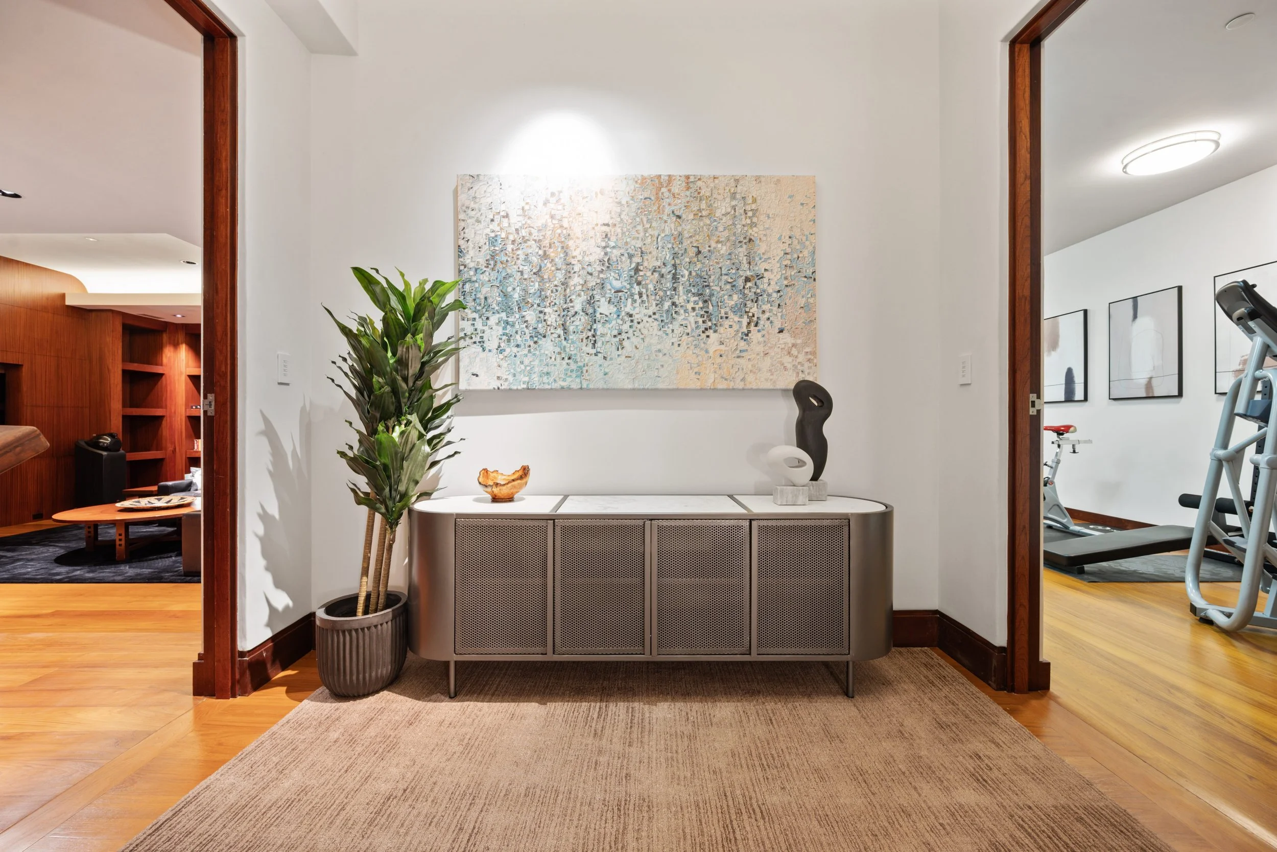 Interior view of a hallway with a modern cabinet, decorative sculptures, a plant, and artwork on the walls leading to a gym area with exercise equipment.