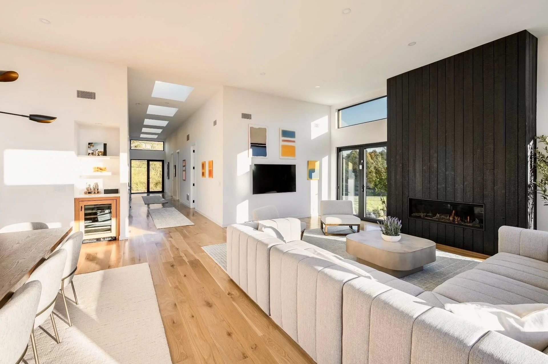 Modern living room with large beige sectional sofa, black accent wall with fireplace, glass sliding doors, and abstract wall art, illuminated by natural light.