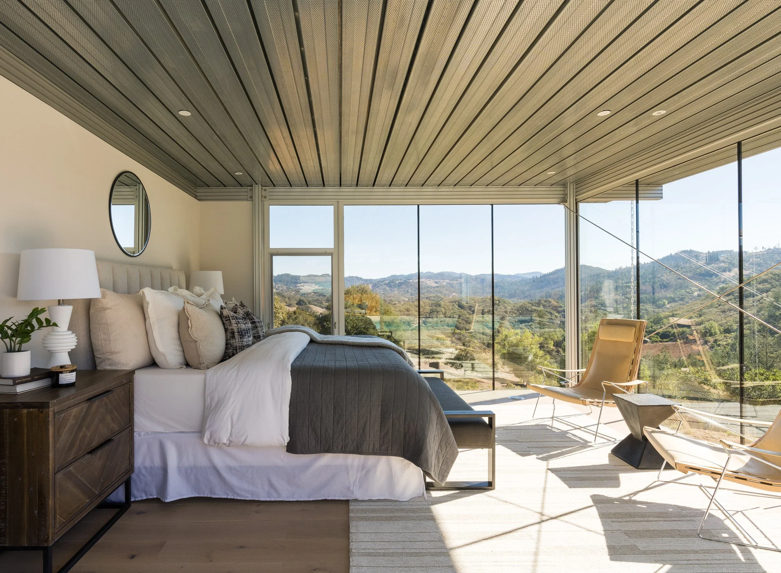 Modern bedroom with a large window overlooking hills, with bed, side table, and chairs.