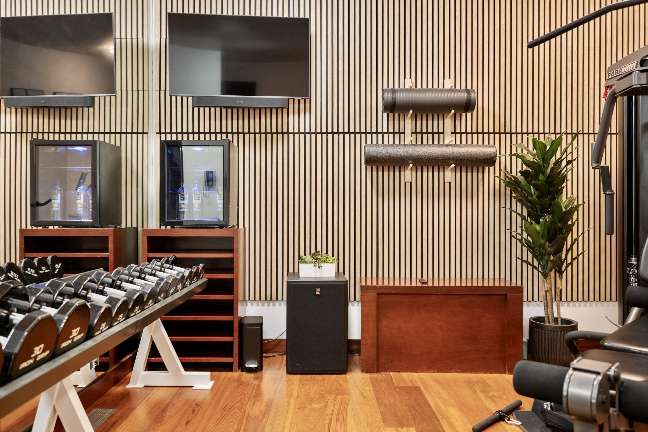 Exercise room with dumbbells, wall-mounted TVs, and foam rollers on a wooden-paneled wall.