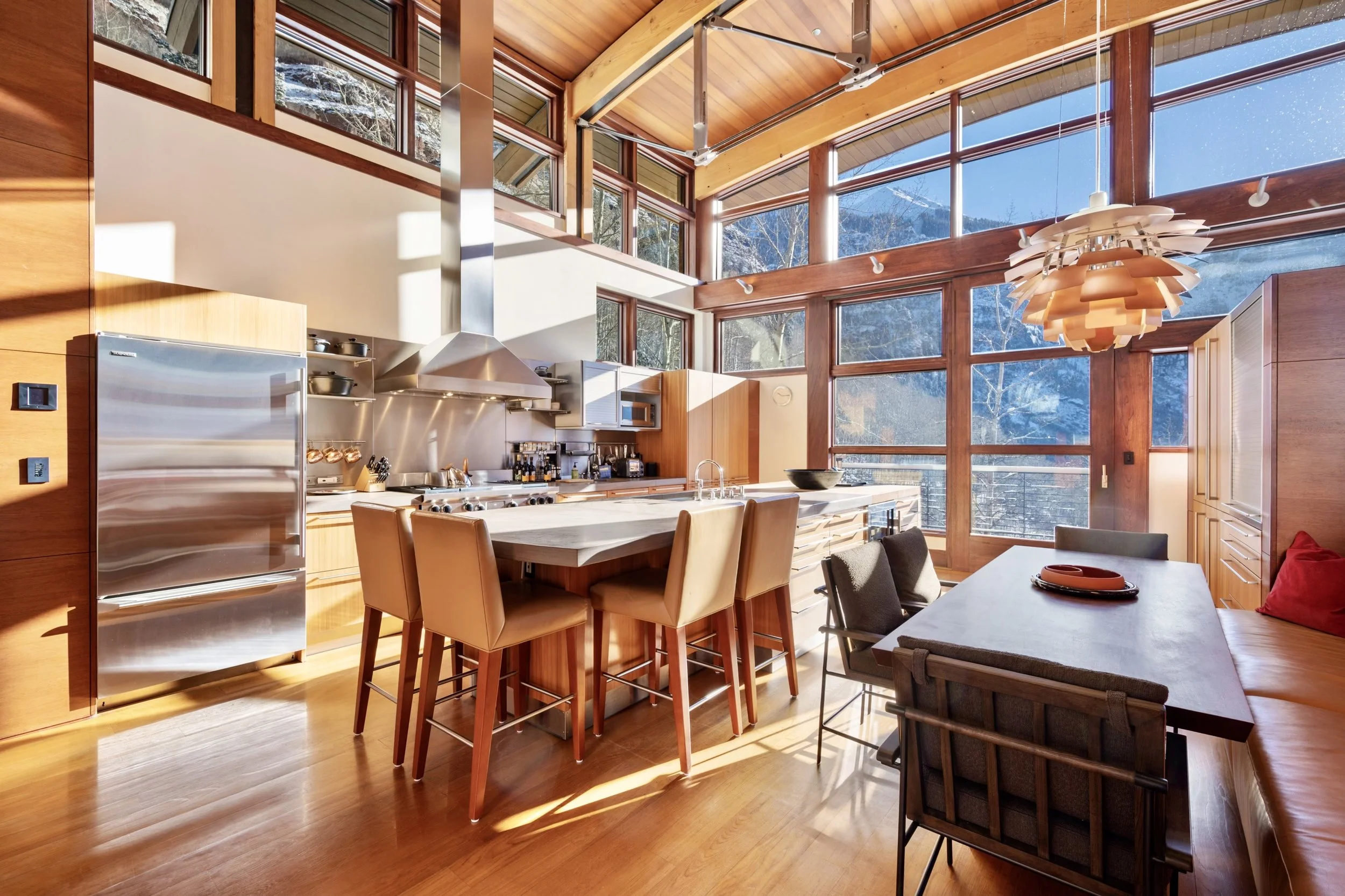 A modern kitchen with large windows showing snow-covered mountains outside. The kitchen features wooden cabinets, a stainless steel refrigerator, a central island, and dining chairs around a table. The ceiling is wood-paneled with a distinctive hangi