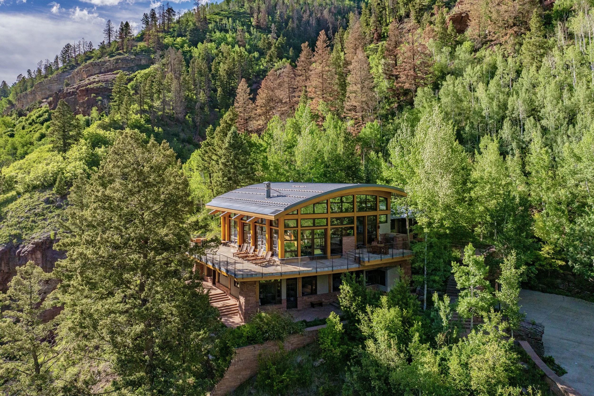 A modern house with a curved roof and extensive glass windows, surrounded by tall green trees on a hillside with a forested mountain in the background.