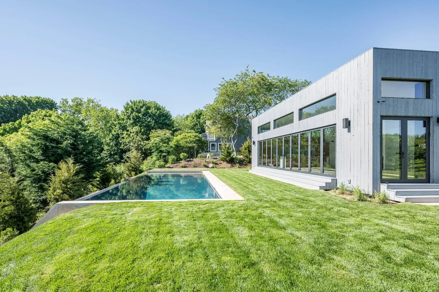 Modern house with large glass windows overlooking a lush green backyard with a swimming pool and trees.