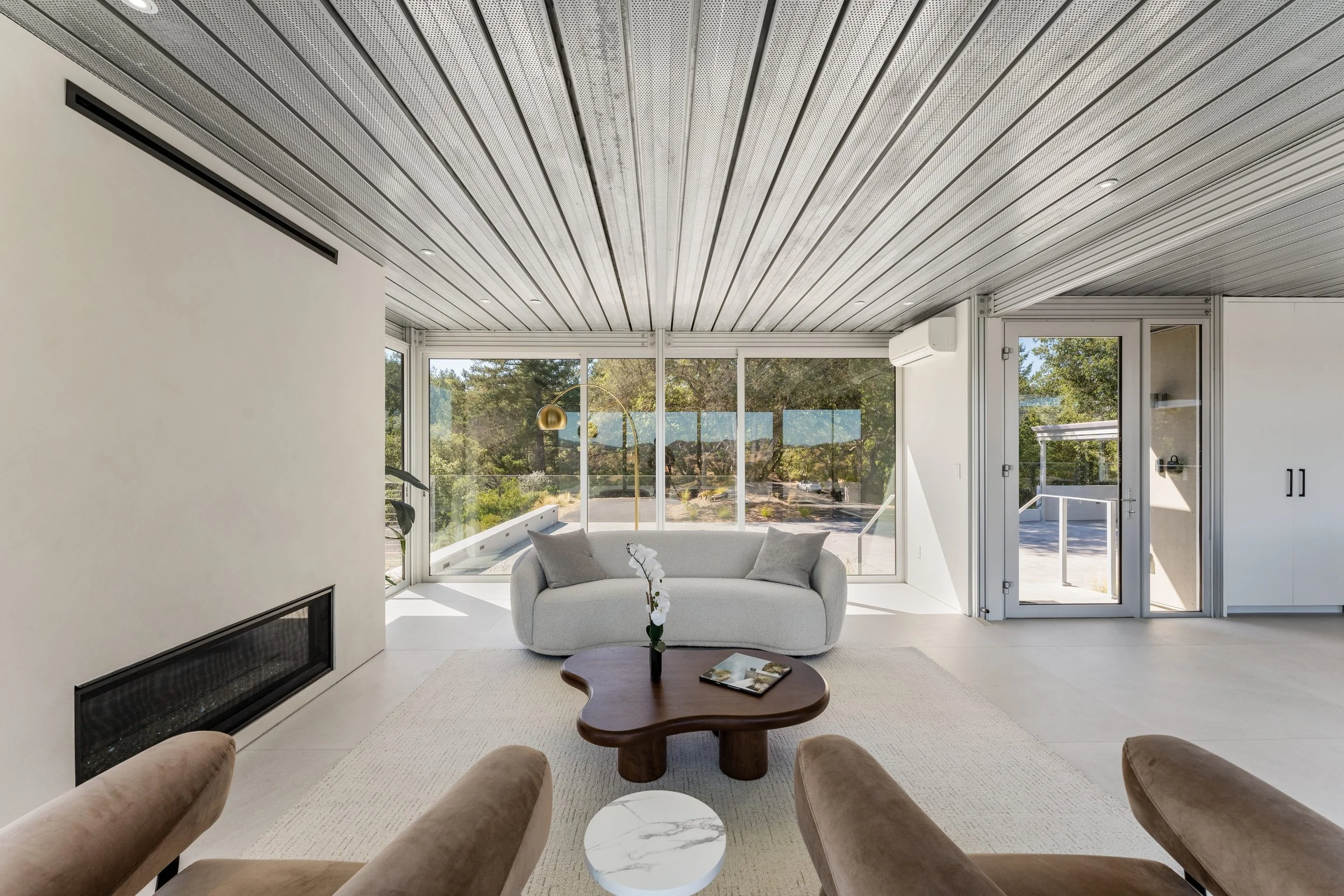 Modern living room with a white couch, a wooden coffee table, large windows revealing trees outside, and a metal ceiling with recessed lighting.