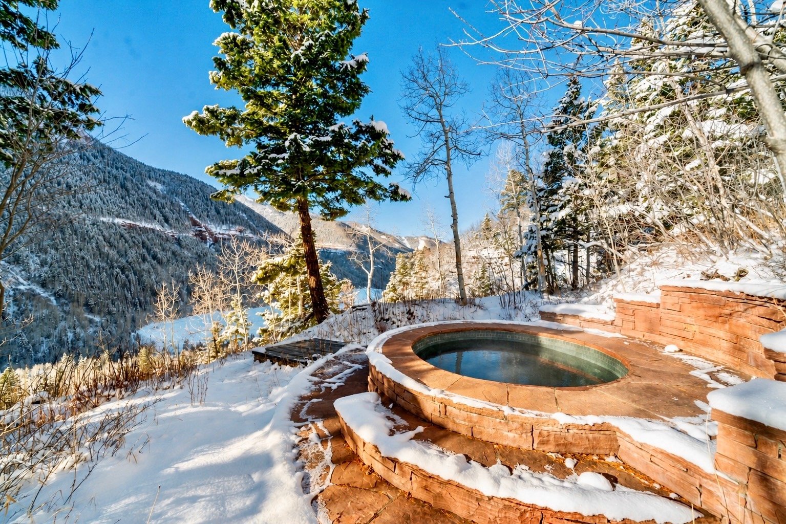 A hot tub surrounded by snow in a mountain landscape with pine trees and a clear blue sky.