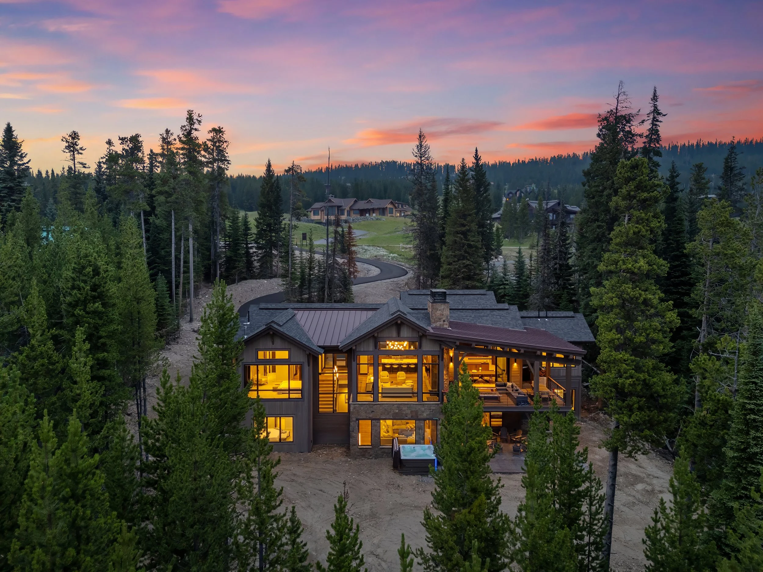 A house with large windows glowing with warm indoor light surrounded by tall pine trees under a colorful sunset sky.
