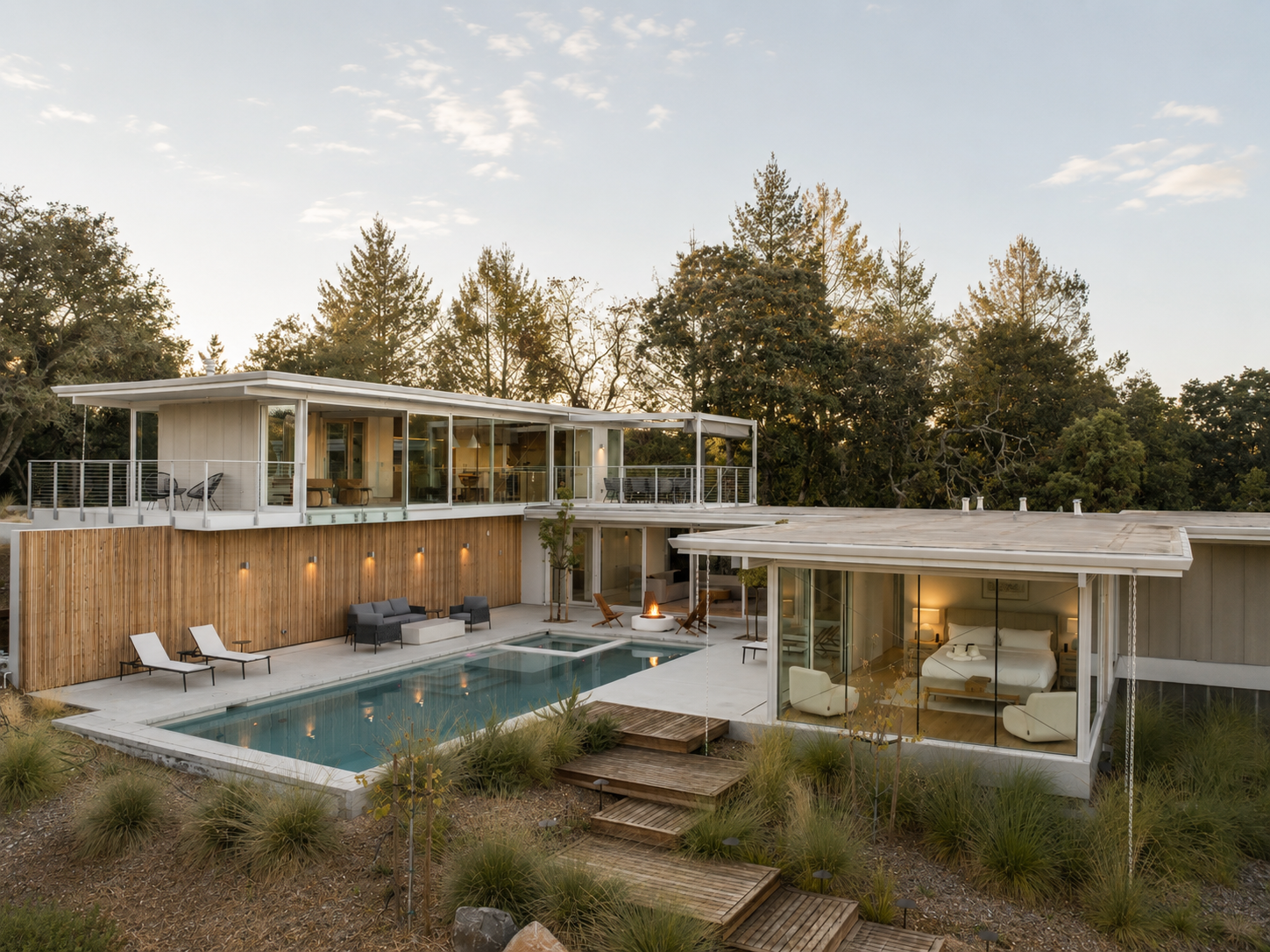 Modern house with large glass windows, outdoor swimming pool, and seating area, surrounded by trees at sunset.