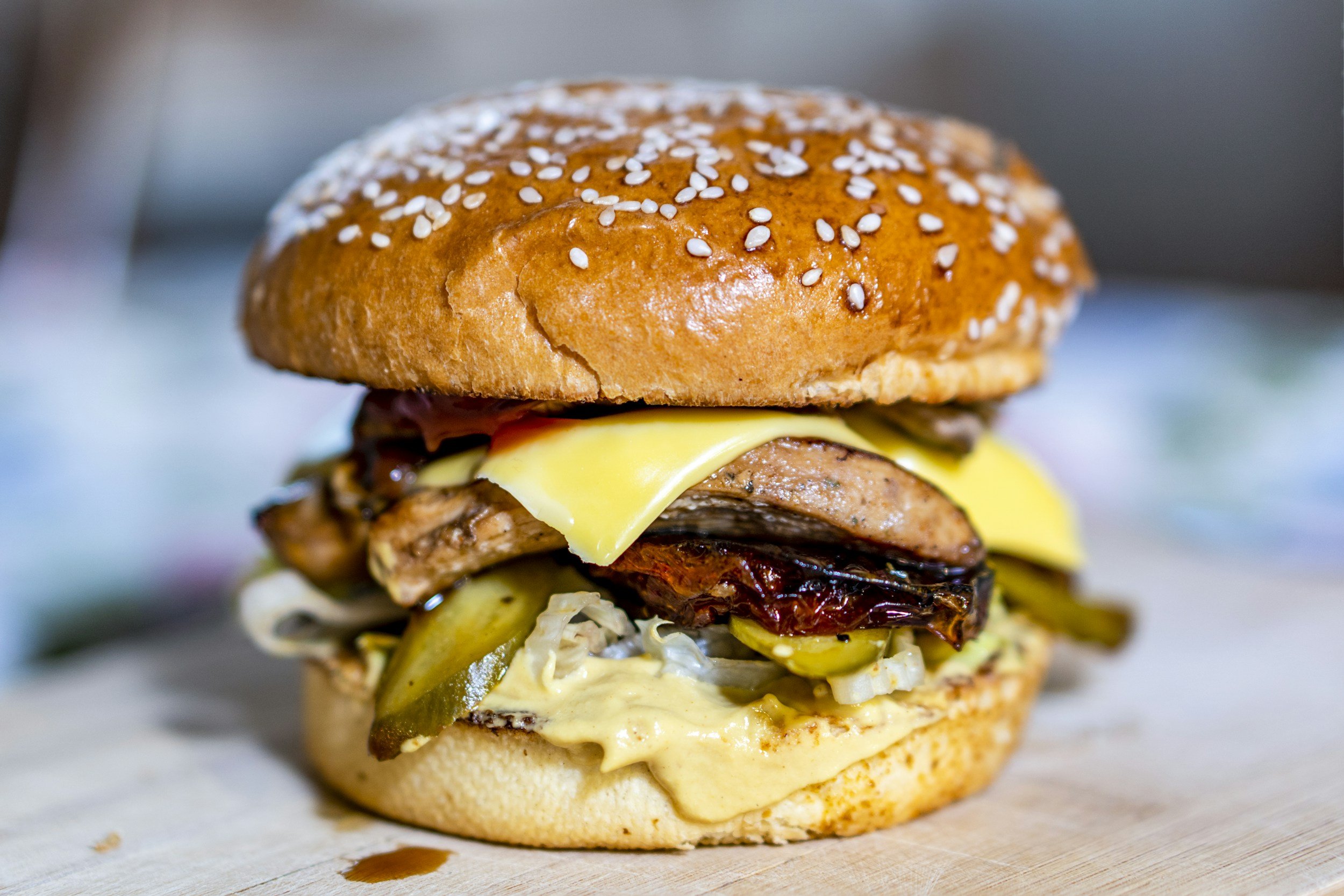 A close-up of a gourmet burger with a sesame seed bun, melted cheese, grilled beef patty, pickles, and various toppings.